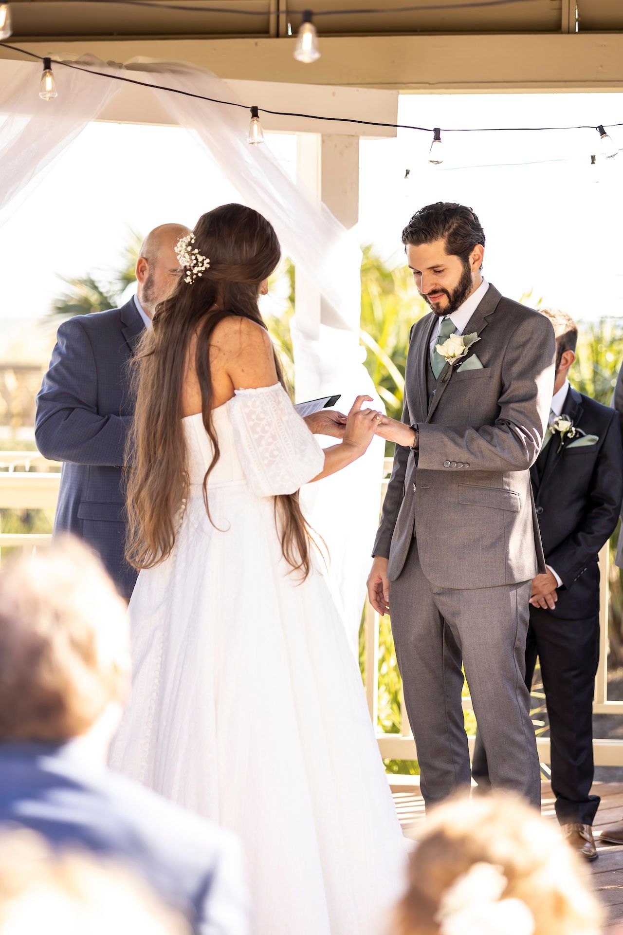 Couple exchanges rings during an outdoor wedding ceremony under a covered pergola with string lights overhead.