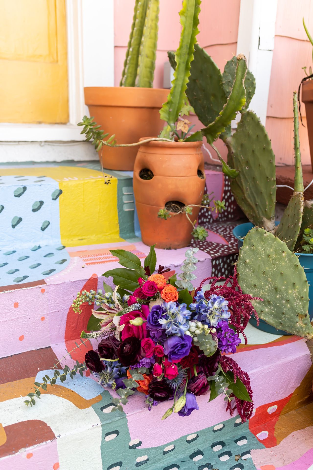 Vibrant floral bouquet with pink and purple blooms sits on colorful geometric outdoor patio near terracotta pots with cacti.
