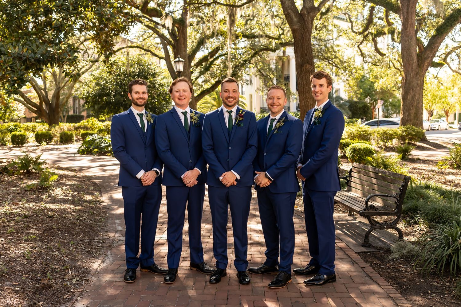 Five people wearing matching navy blue suits standing together on a path under moss-draped oak trees in a garden setting.