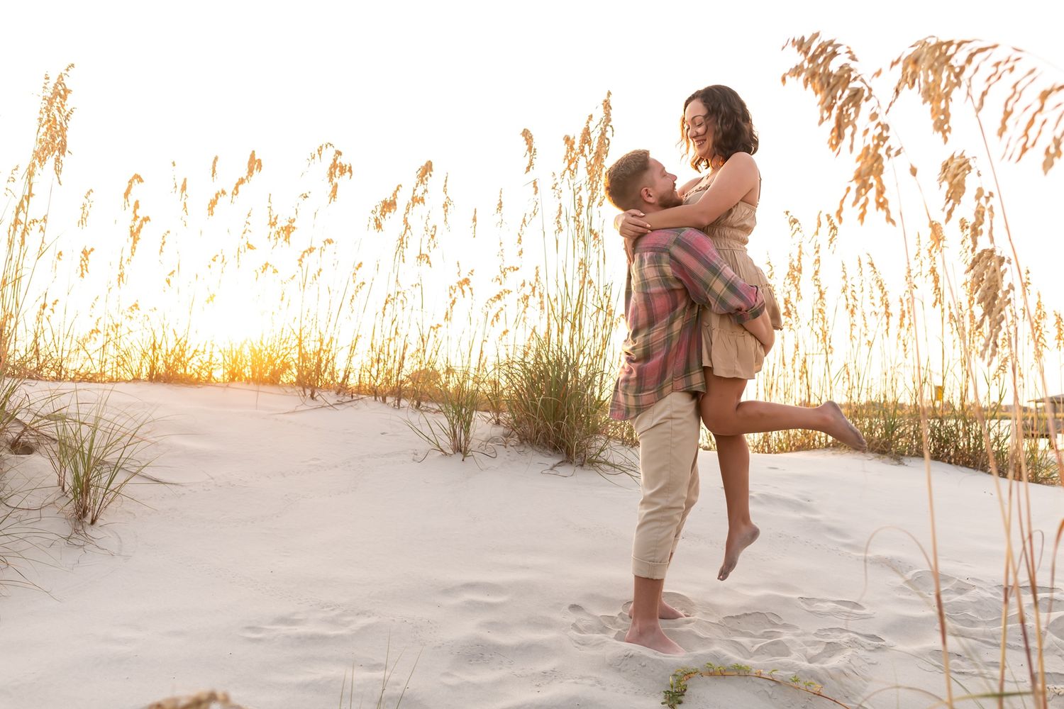 Couple embraces on sandy beach at sunset surrounded by golden sea grass.