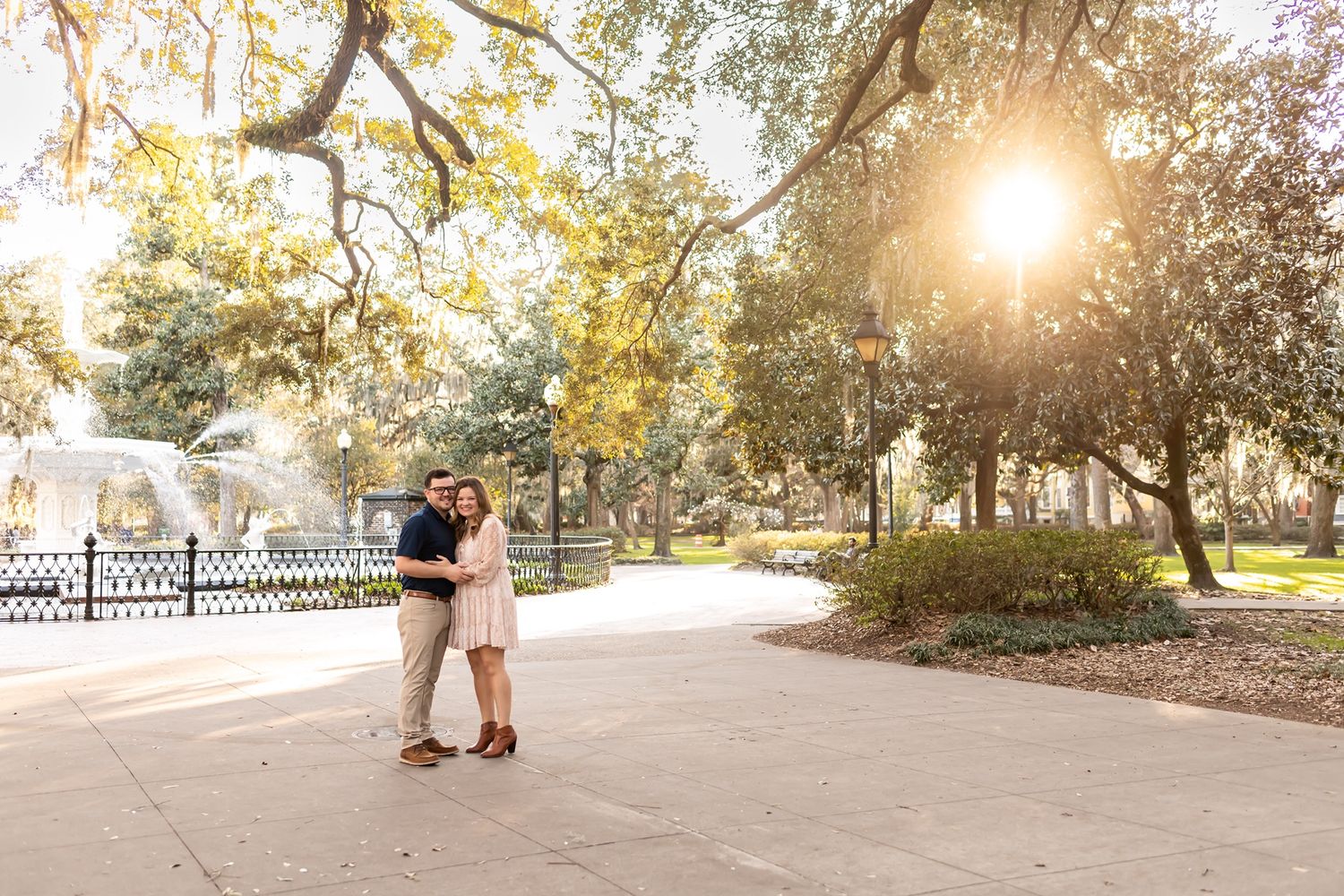 A couple shares a romantic moment at sunset in a picturesque park setting with golden sunlight streaming through trees.