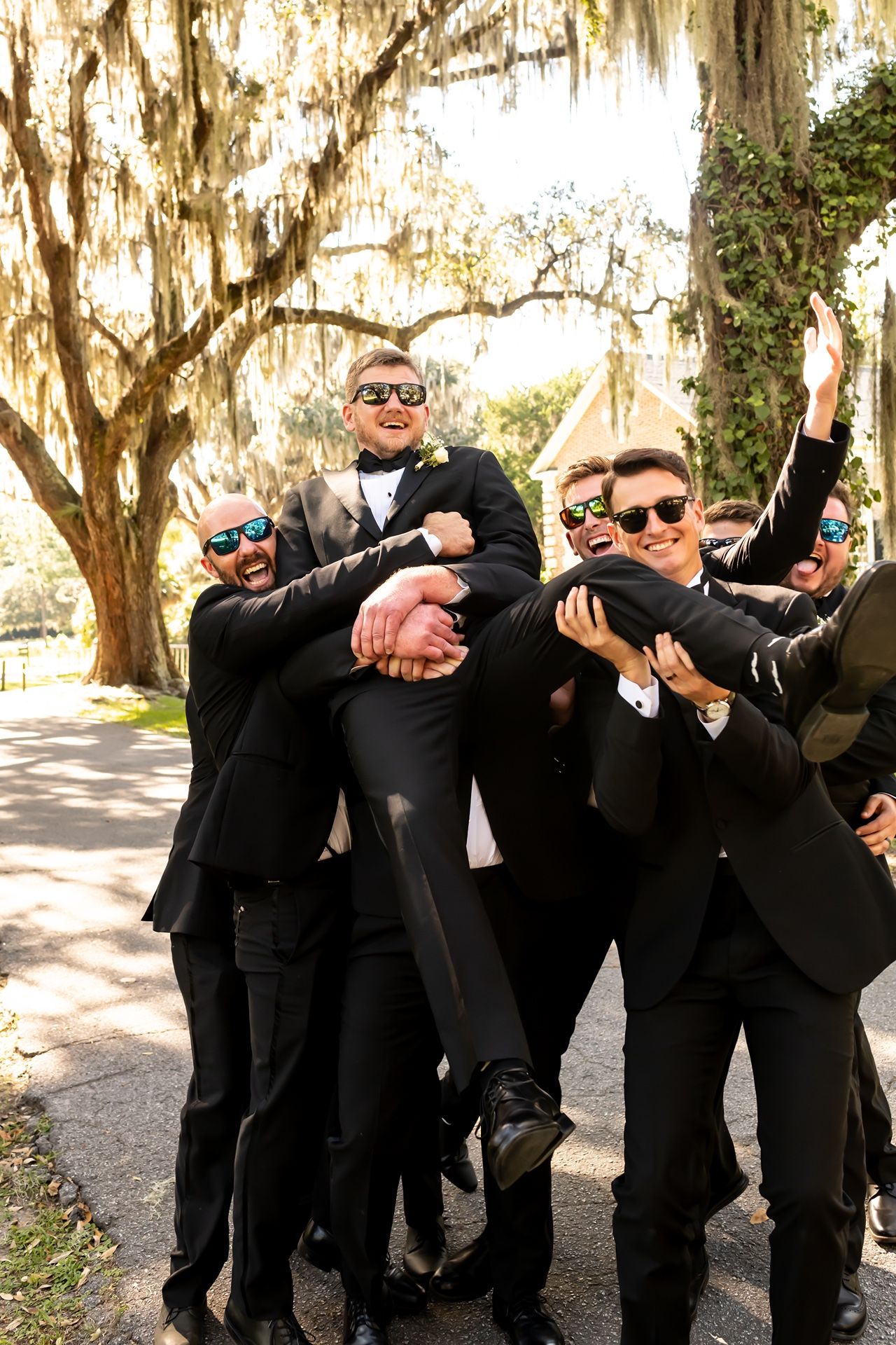 Groomsmen in black suits pose playfully making secret agent gestures on a tree-lined path.