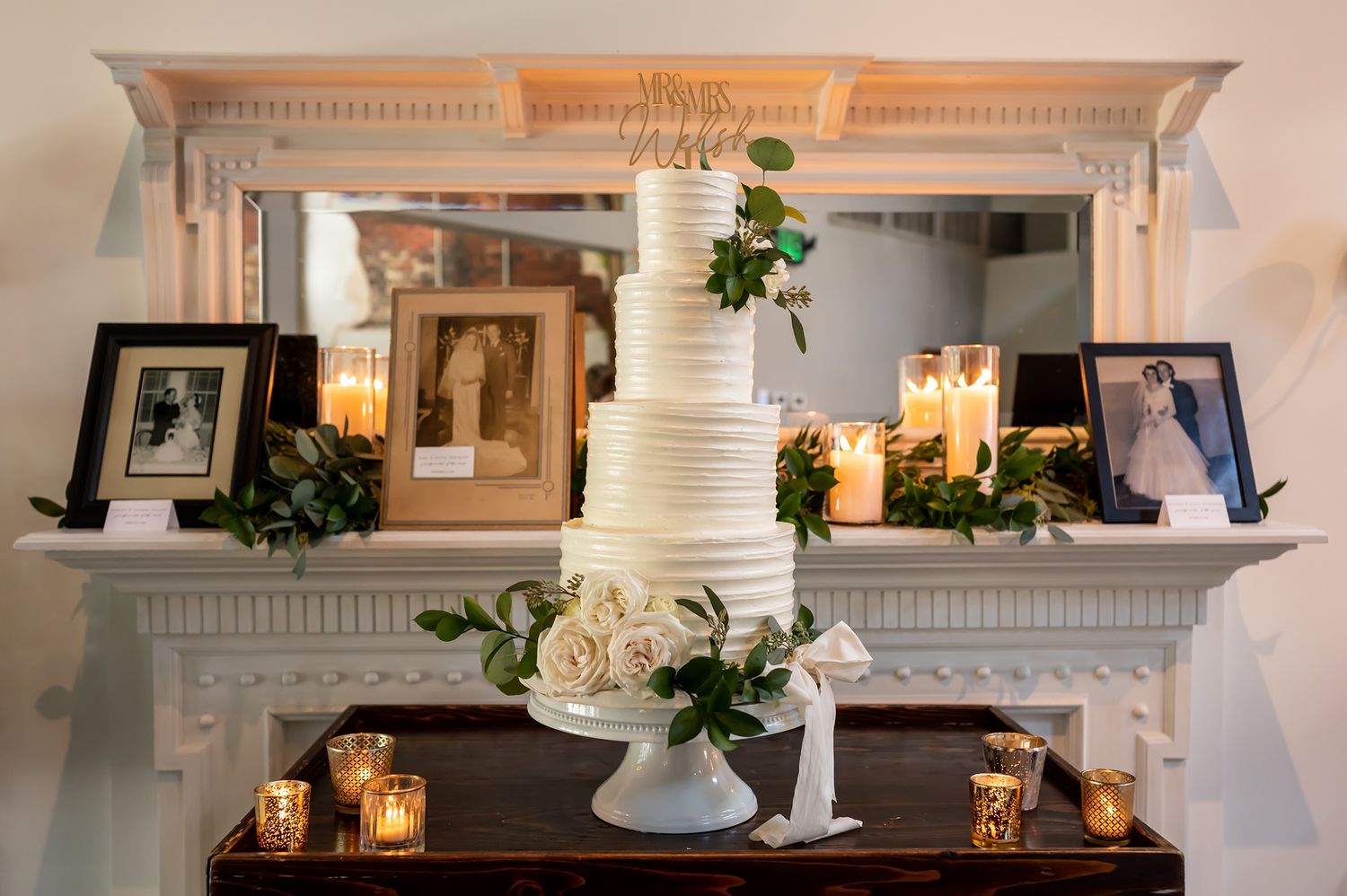 White fireplace mantel decorated with glowing candles, greenery and rustic lanterns creates a cozy holiday atmosphere.