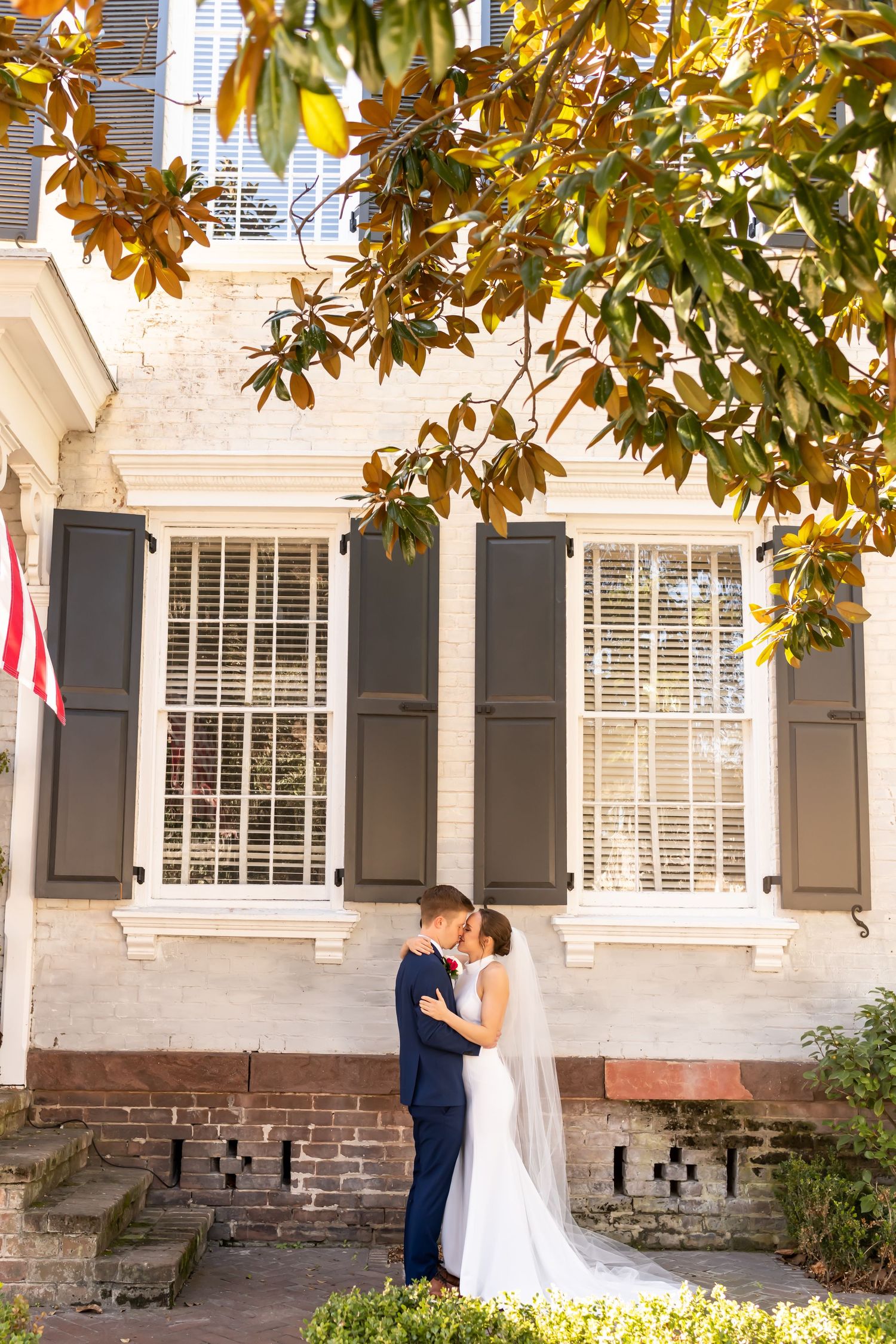 Wedding couple shares romantic moment under golden magnolia tree canopy on southern plantation veranda.
