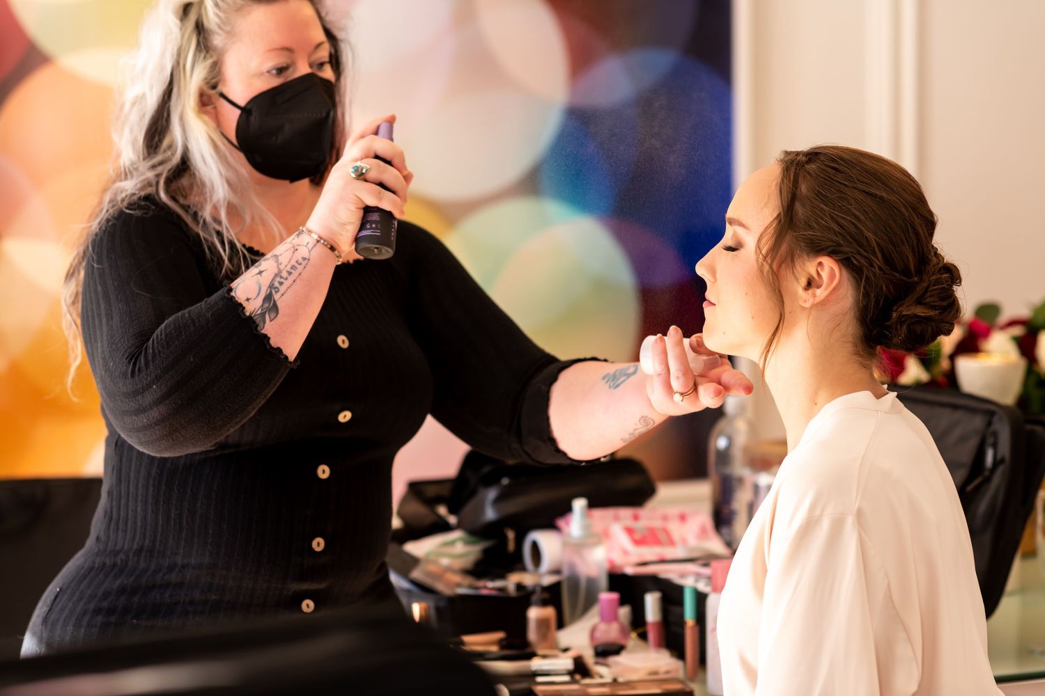 A makeup artist wearing a black face mask applies cosmetics to a client in a colorfullylit setting.