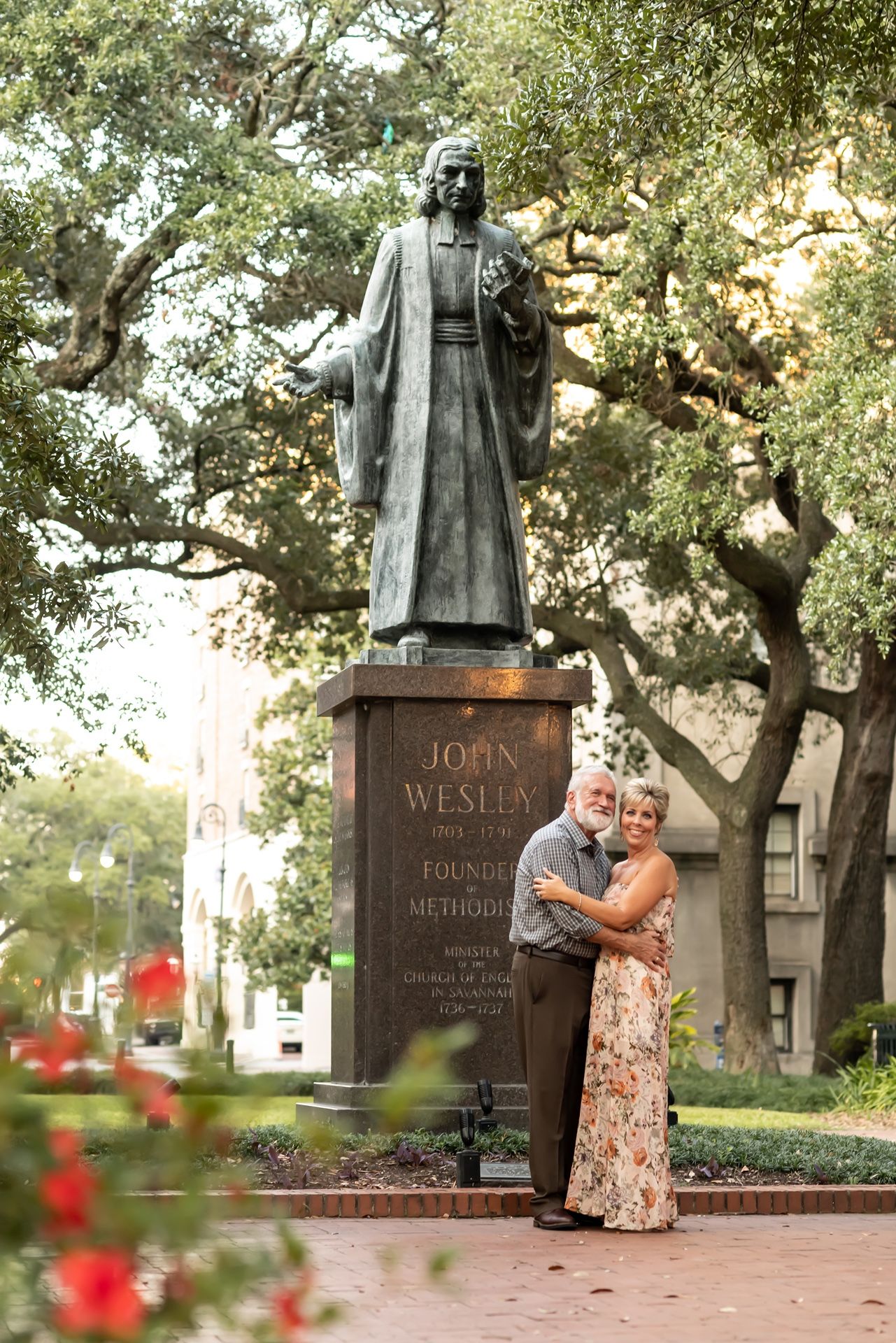A couple embraces near a statue of John Wesley surrounded by trees in a historic square.