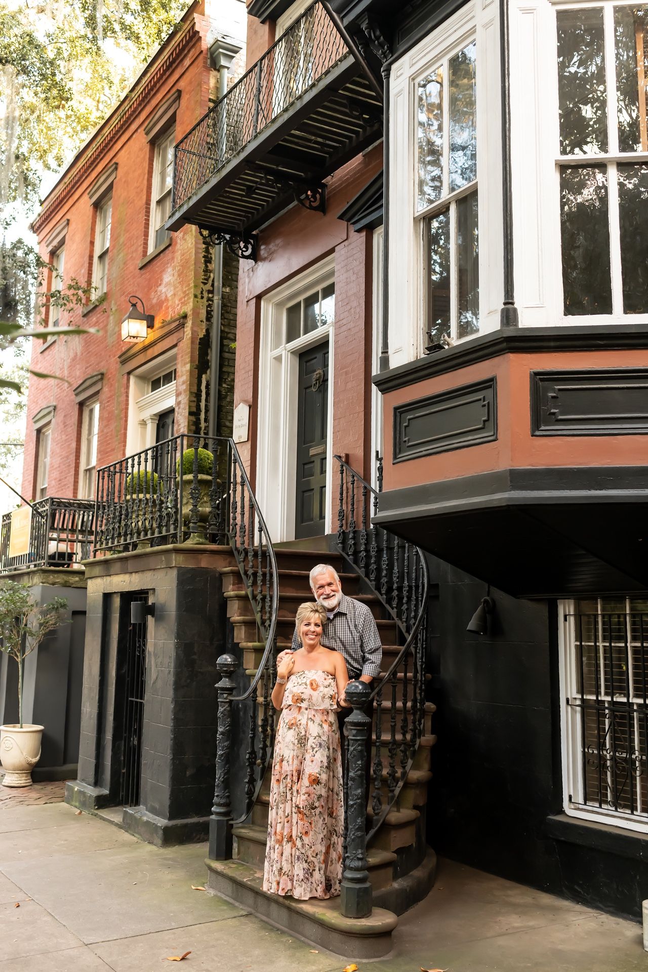 A couple stands on ornate iron steps outside a historic brownstone building with decorative architectural details.