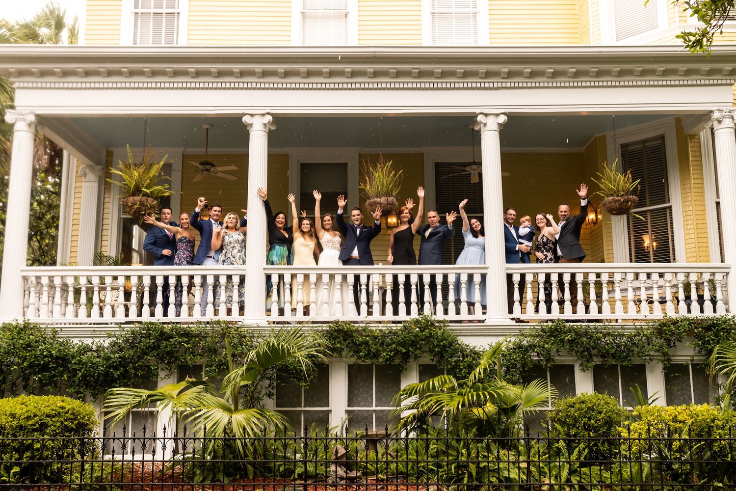 Large group gathered on an elegant white-columned Southern mansion porch surrounded by lush greenery and gardens.