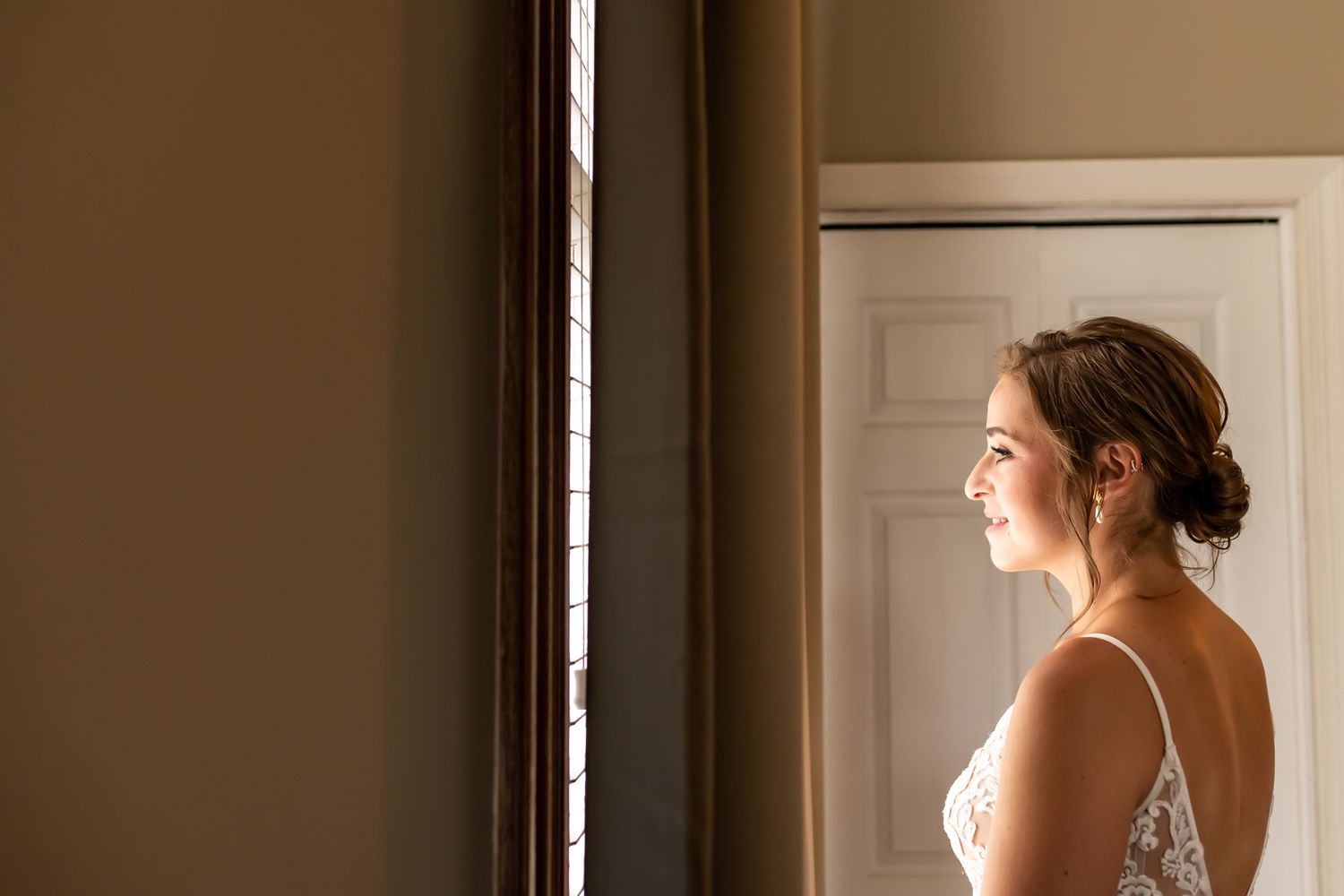 A bride in a white wedding dress stands in soft window light while gazing outside on her wedding day.