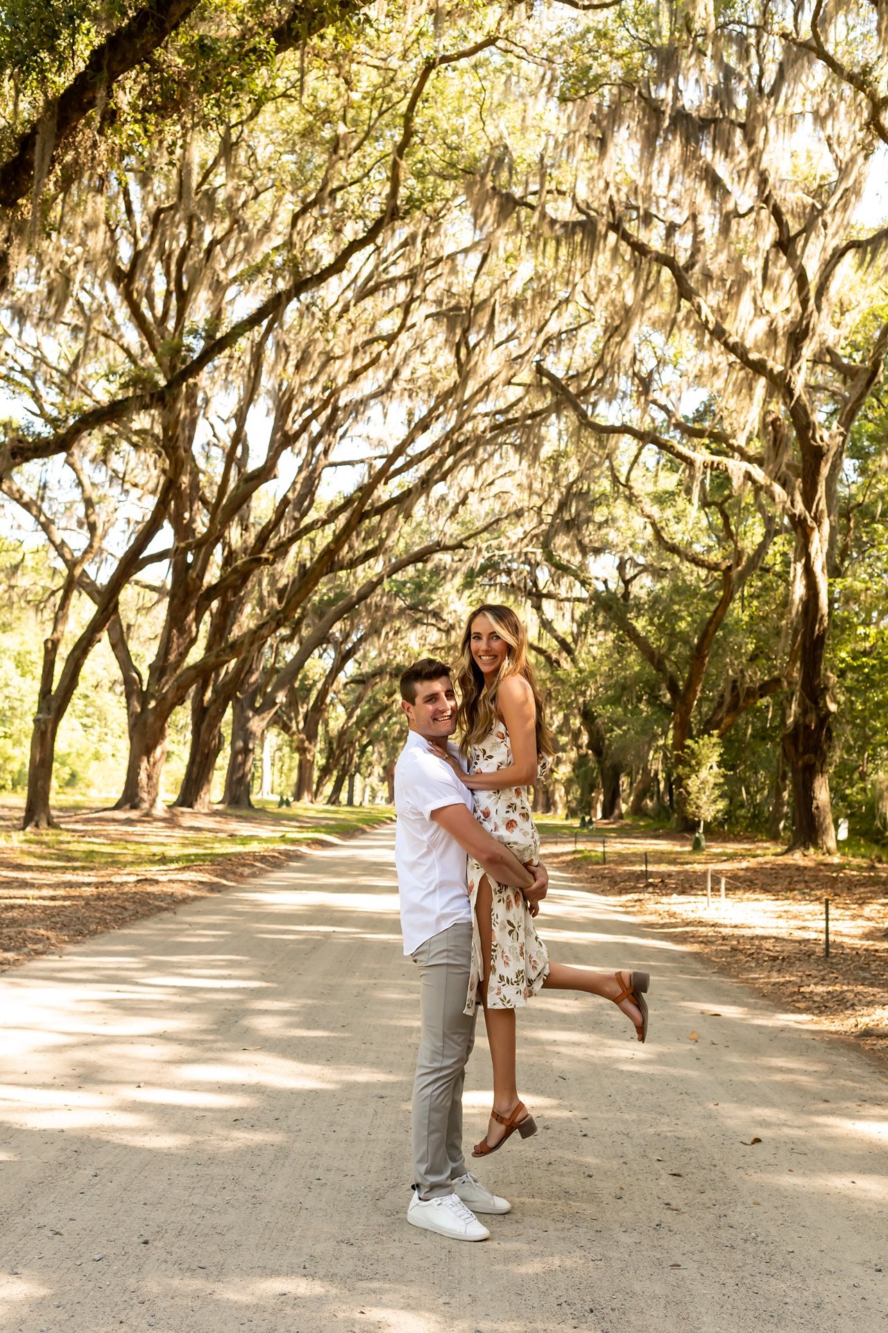 A romantic moment captured beneath majestic Spanish moss-draped oak trees along a dirt path on a sunny afternoon.