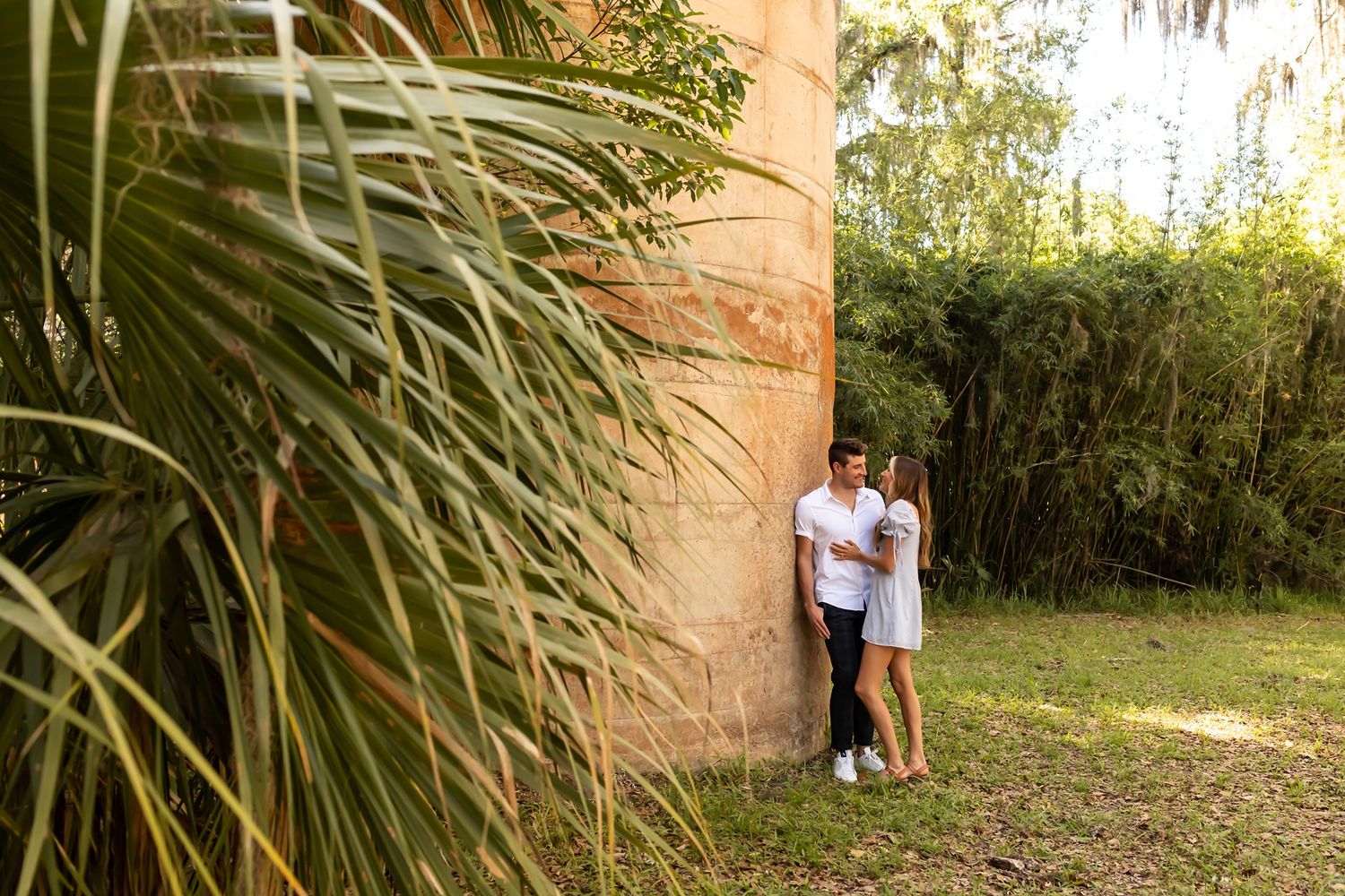 Two figures embrace beside a stone wall surrounded by tropical plants and grass in an outdoor garden setting.