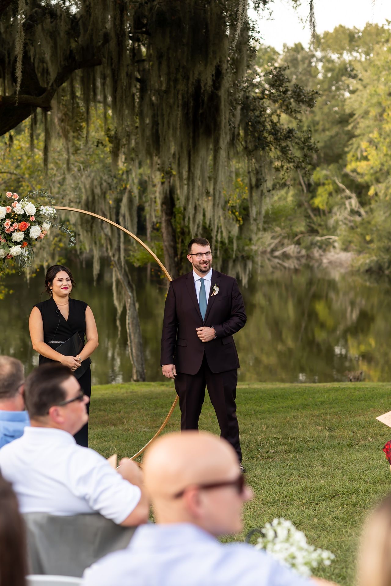 An outdoor wedding ceremony beside a tranquil lake with Spanish moss hanging from trees in the background.