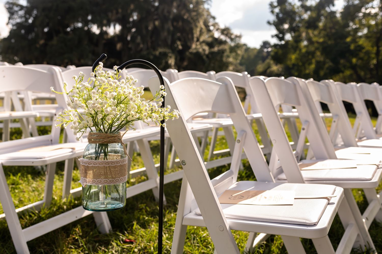 White folding chairs lined up for an outdoor wedding ceremony with mason jar floral centerpieces decorating the aisle.