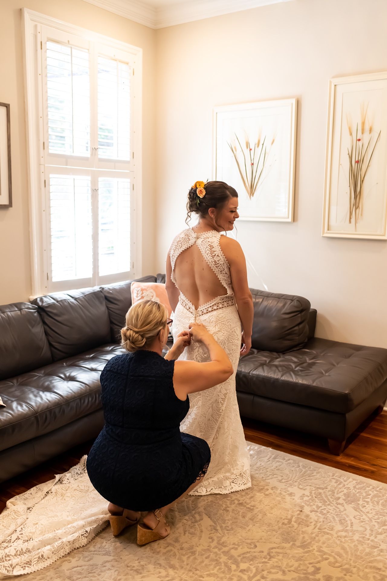 Someone helps adjust the train of an elegant lace wedding dress with an open back design in a living room setting.