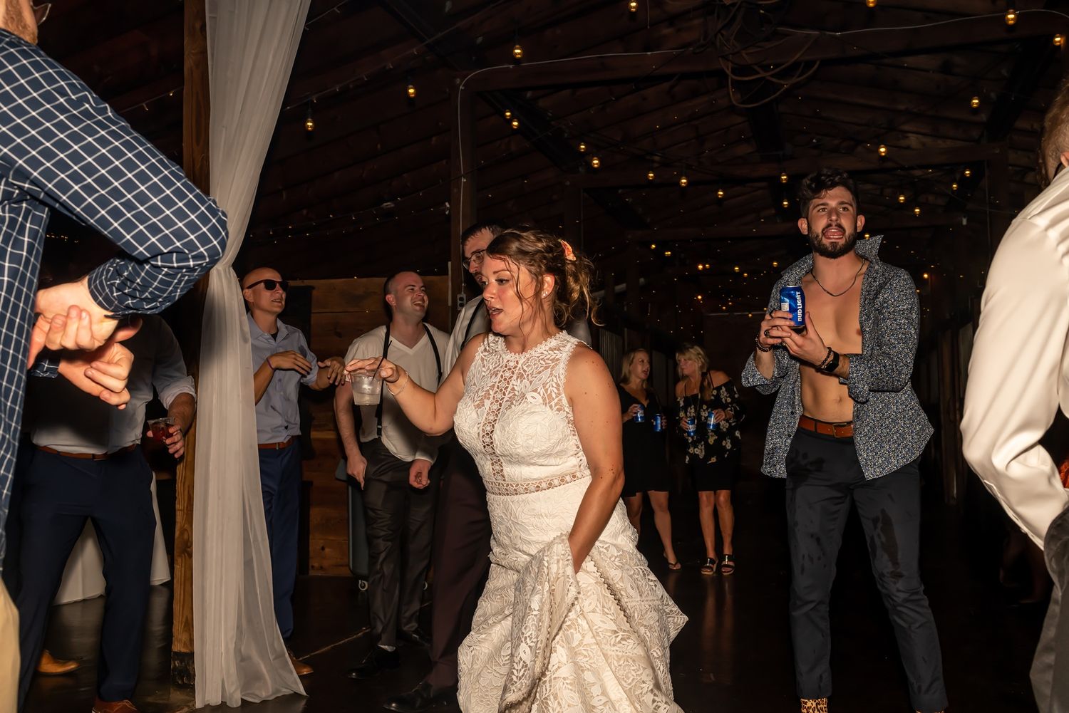 Wedding guests dancing and celebrating on a dimly lit dance floor surrounded by white drapery and string lights.