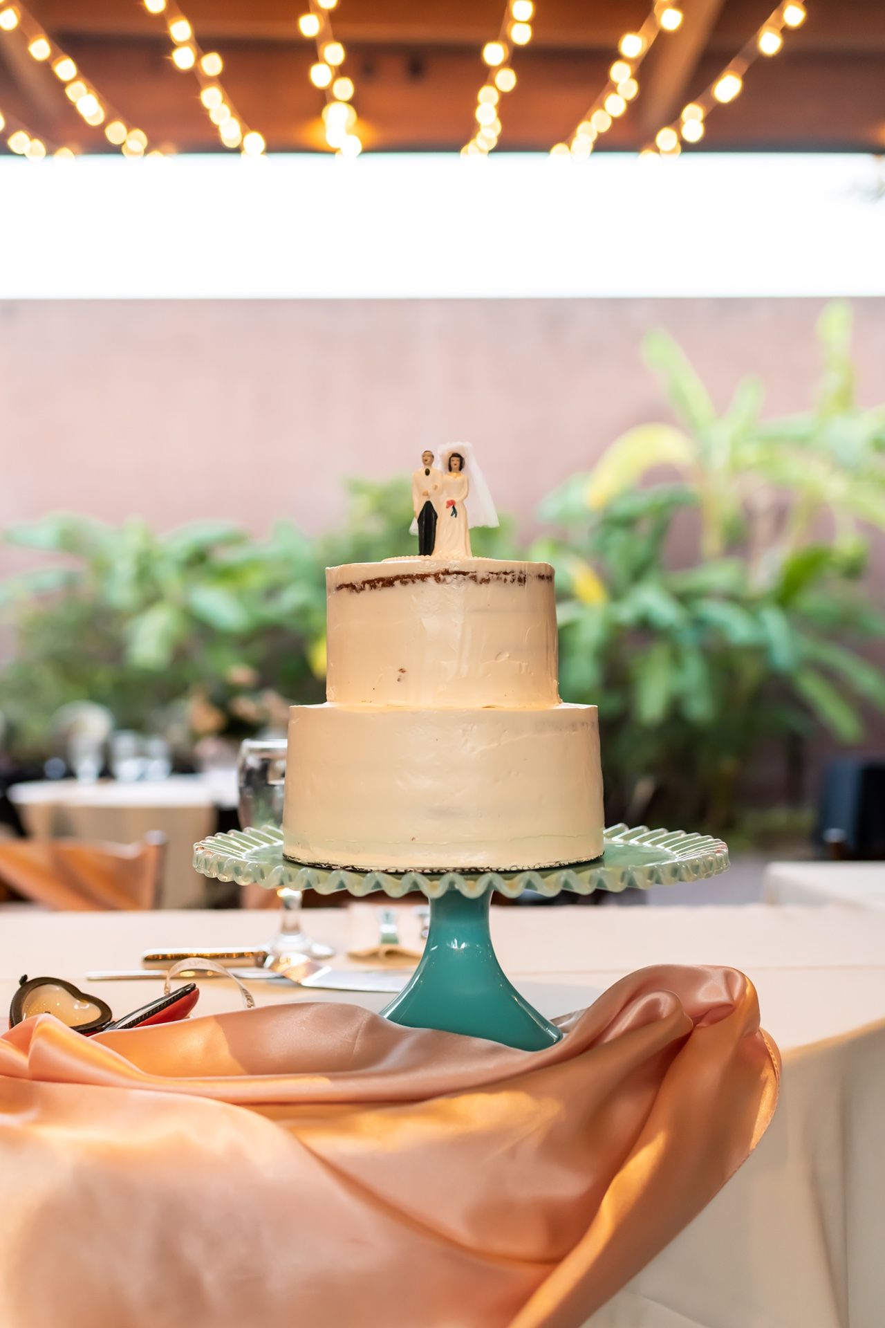 Two-tiered white wedding cake with bride and groom topper displayed on turquoise pedestal with peach tablecloth and twinkle lights.