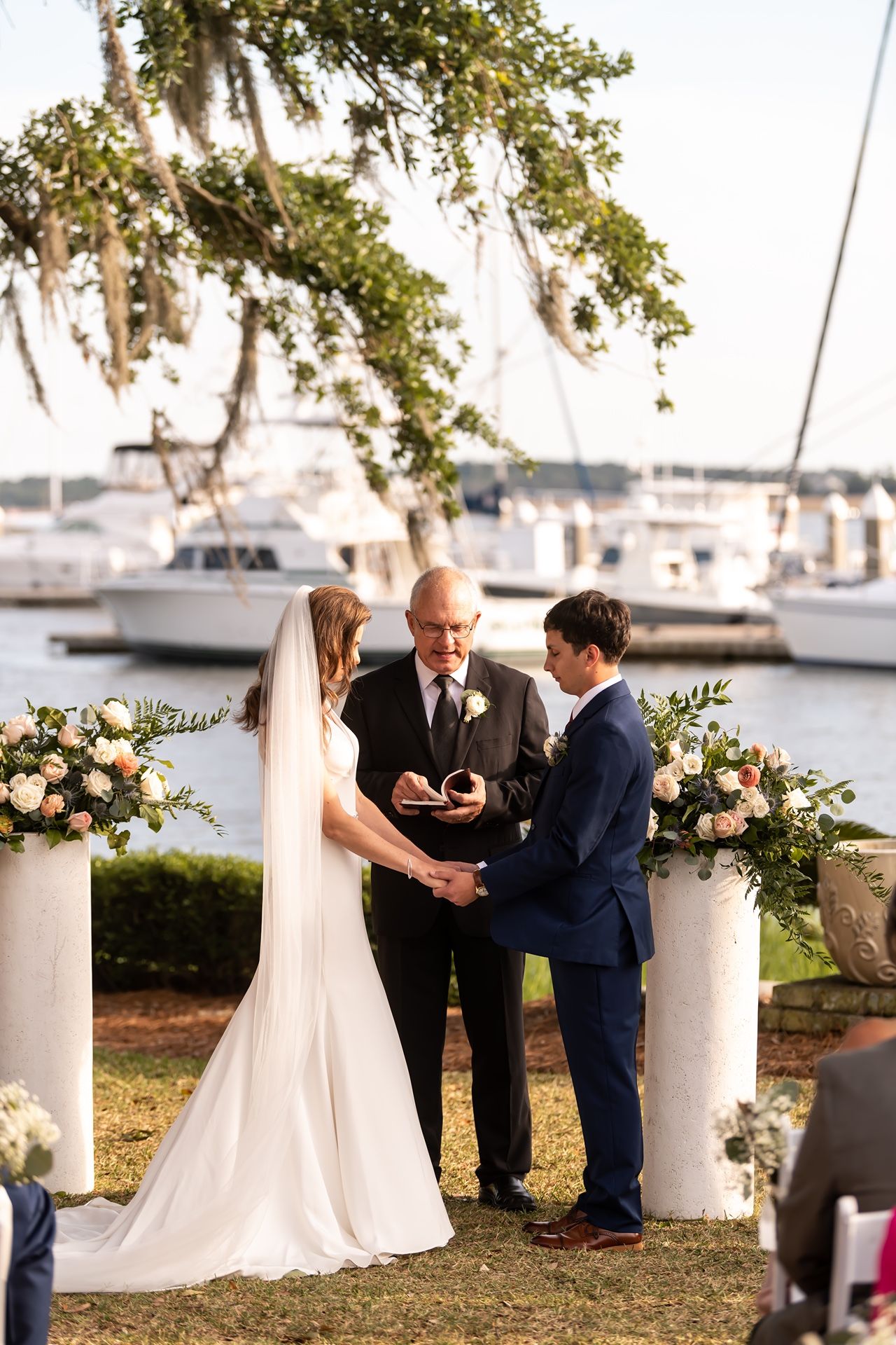 Wedding ceremony taking place at waterfront venue with white columns and floral decorations.