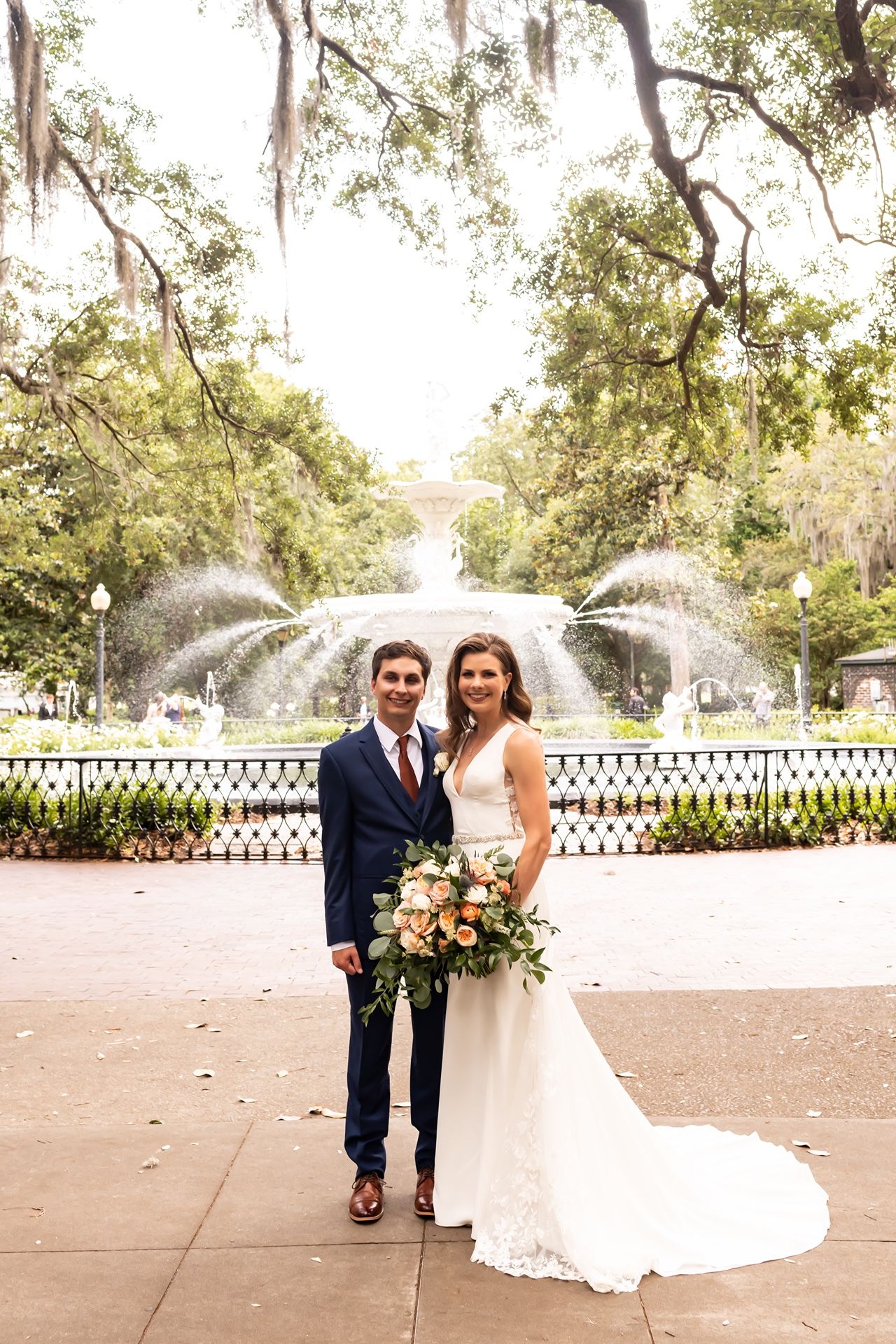 Newlywed couple poses in front of ornate fountain with Spanish moss draped trees in Savannah setting.