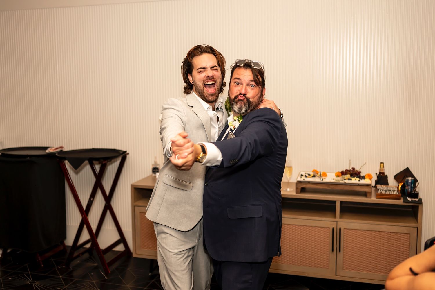 Two people in suits laughing and embracing while standing near a piano and cabinet in a room.