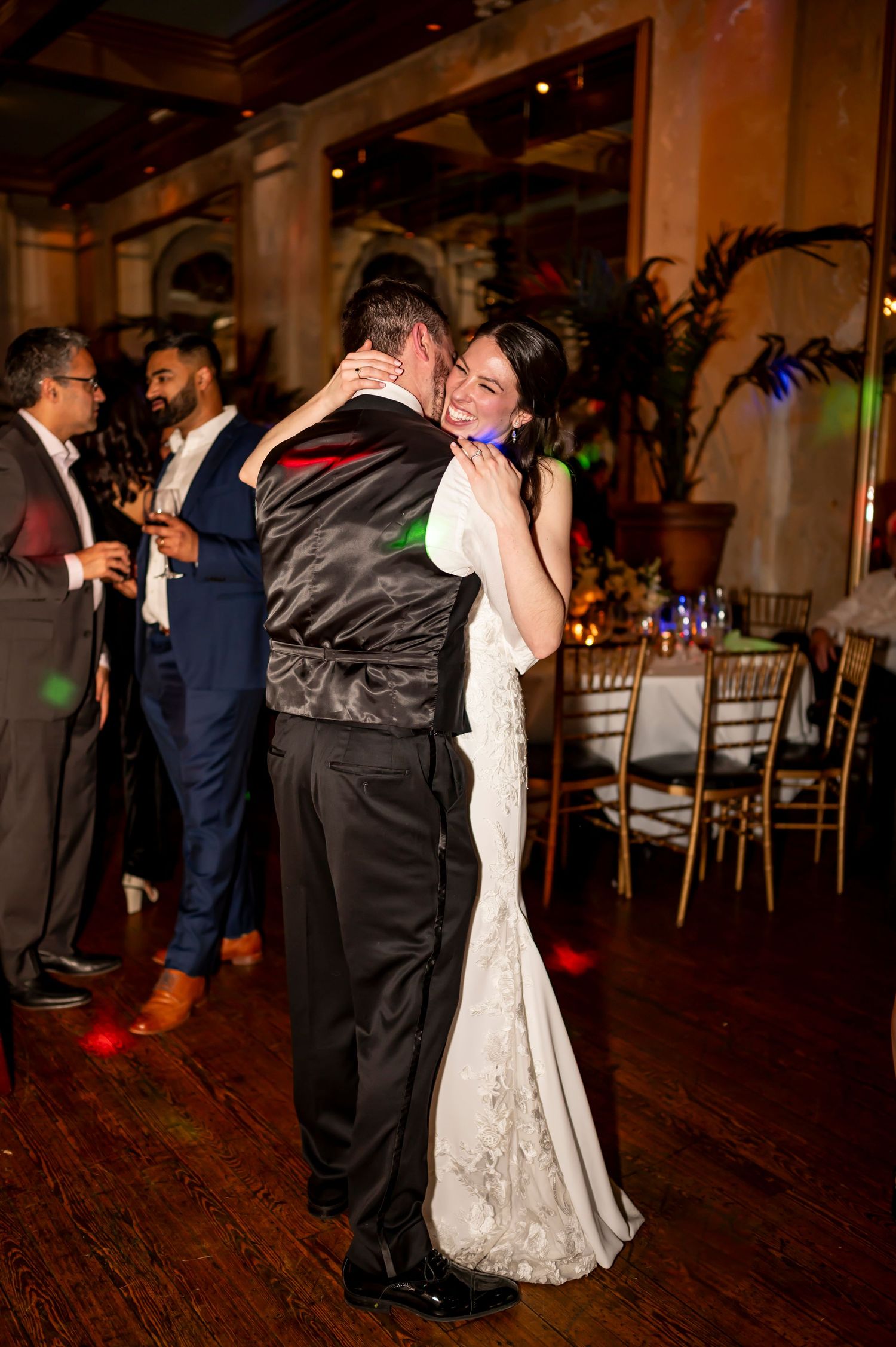 A couple shares an intimate dance together at their wedding reception on a wooden floor with guests in the background.