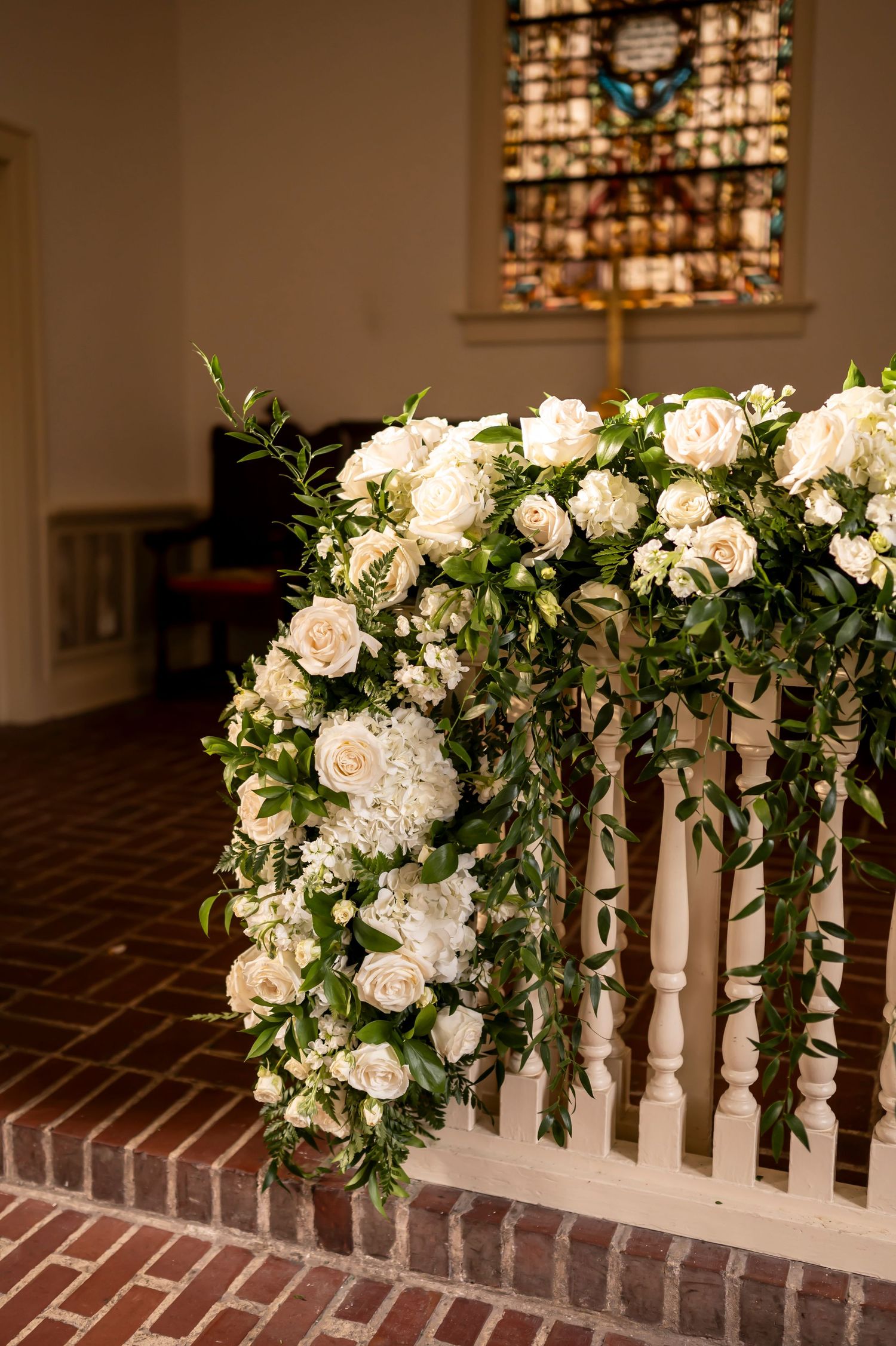White roses and greenery elegantly cascade over a white railing in a church with a stained glass window in background.