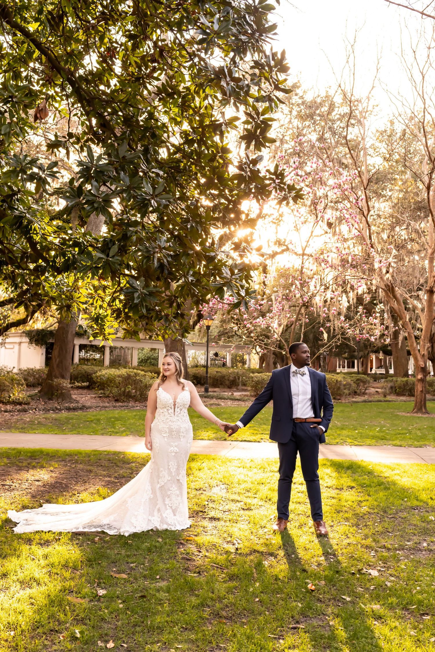 A couple holds hands in a sun-drenched garden, the bride wearing a fitted lace gown and groom in classic black tuxedo.