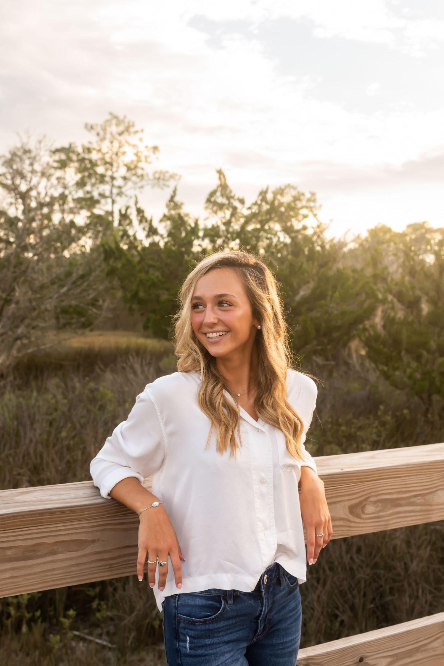 Person in white button-up shirt and jeans leaning against wooden railing at sunset with trees in background.