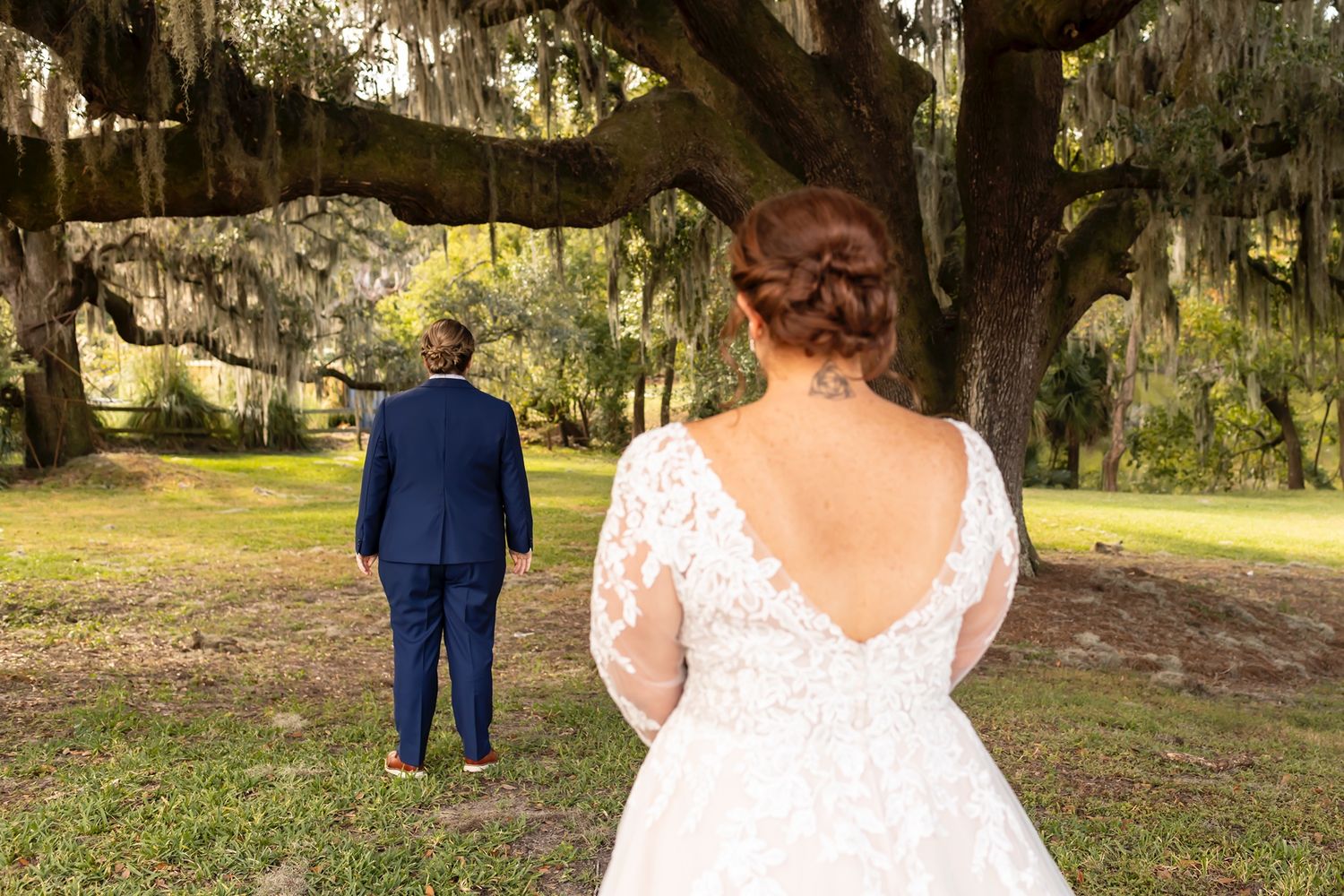 A wedding couple stands apart during their first look moment in a garden with Spanish moss draped trees.