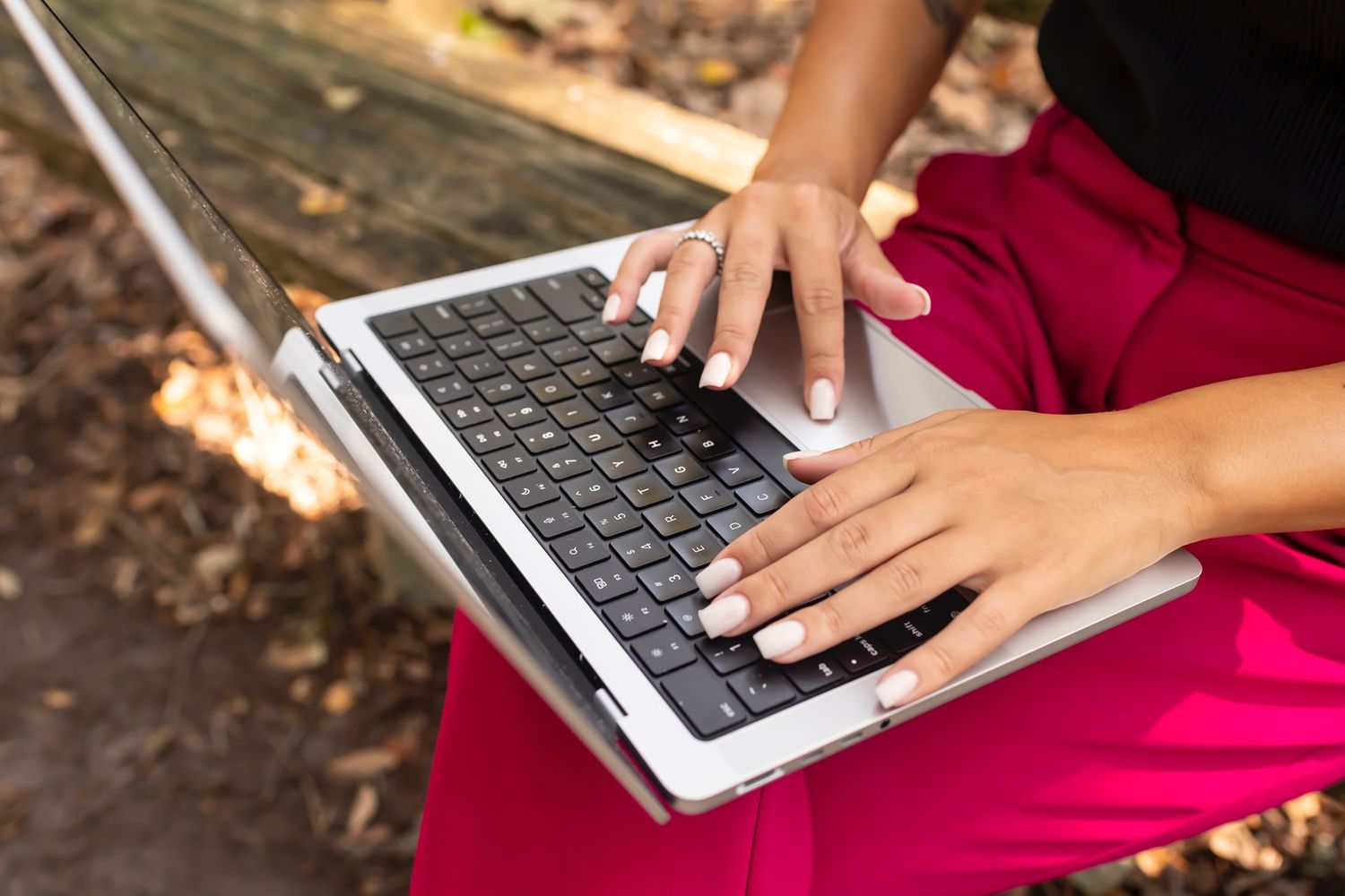 Hands typing on silver laptop keyboard while wearing red skirt outdoors in natural setting.