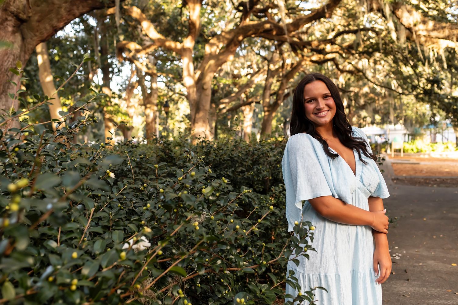 Person in white dress stands smiling along tree-lined pathway with golden sunlight filtering through branches.