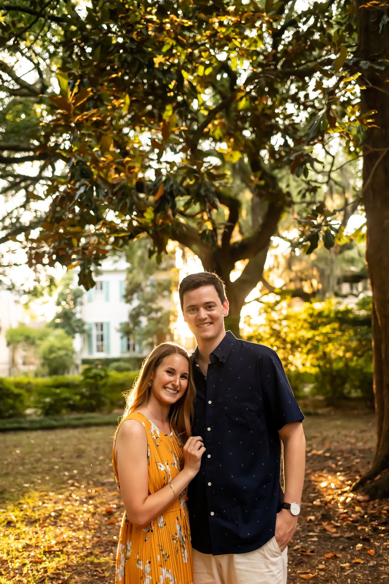 A couple stands together on fallen autumn leaves during golden hour with sunlight filtering through trees.