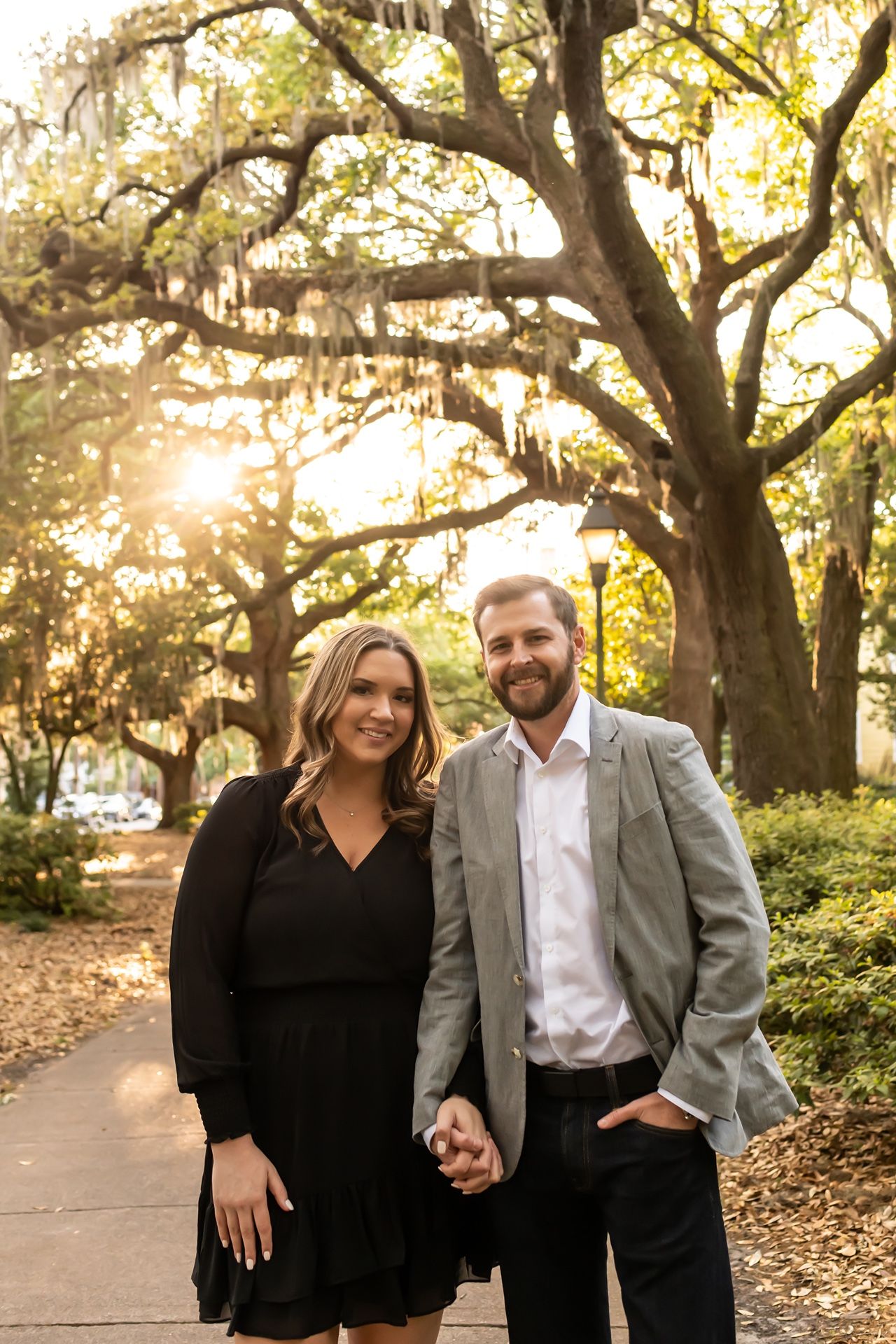A couple poses together on a pathway beneath Spanish moss-draped oak trees at sunset in a romantic outdoor setting.