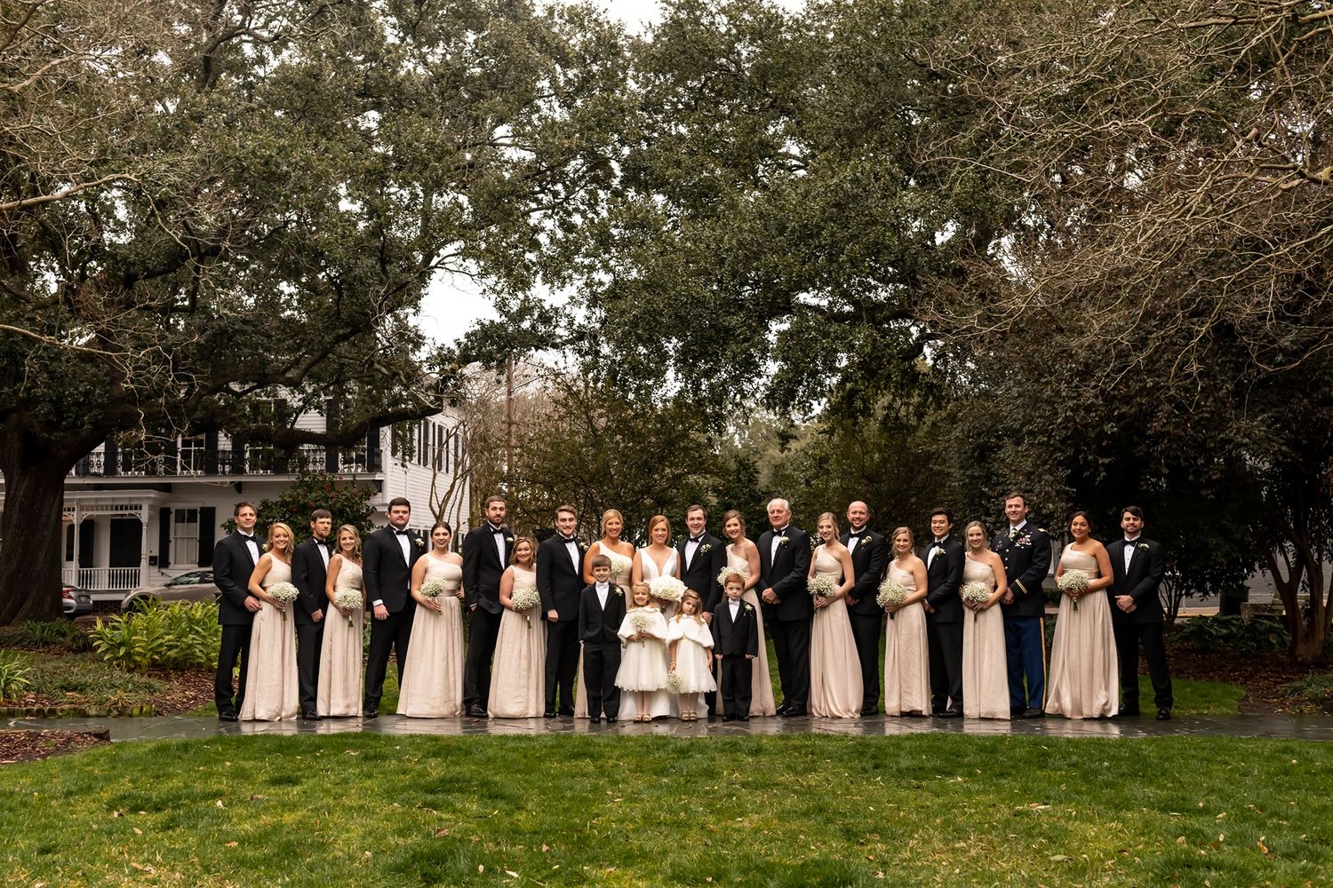 Wedding party in pink dresses and black suits stand in line on grassy lawn beneath large trees and historic building.