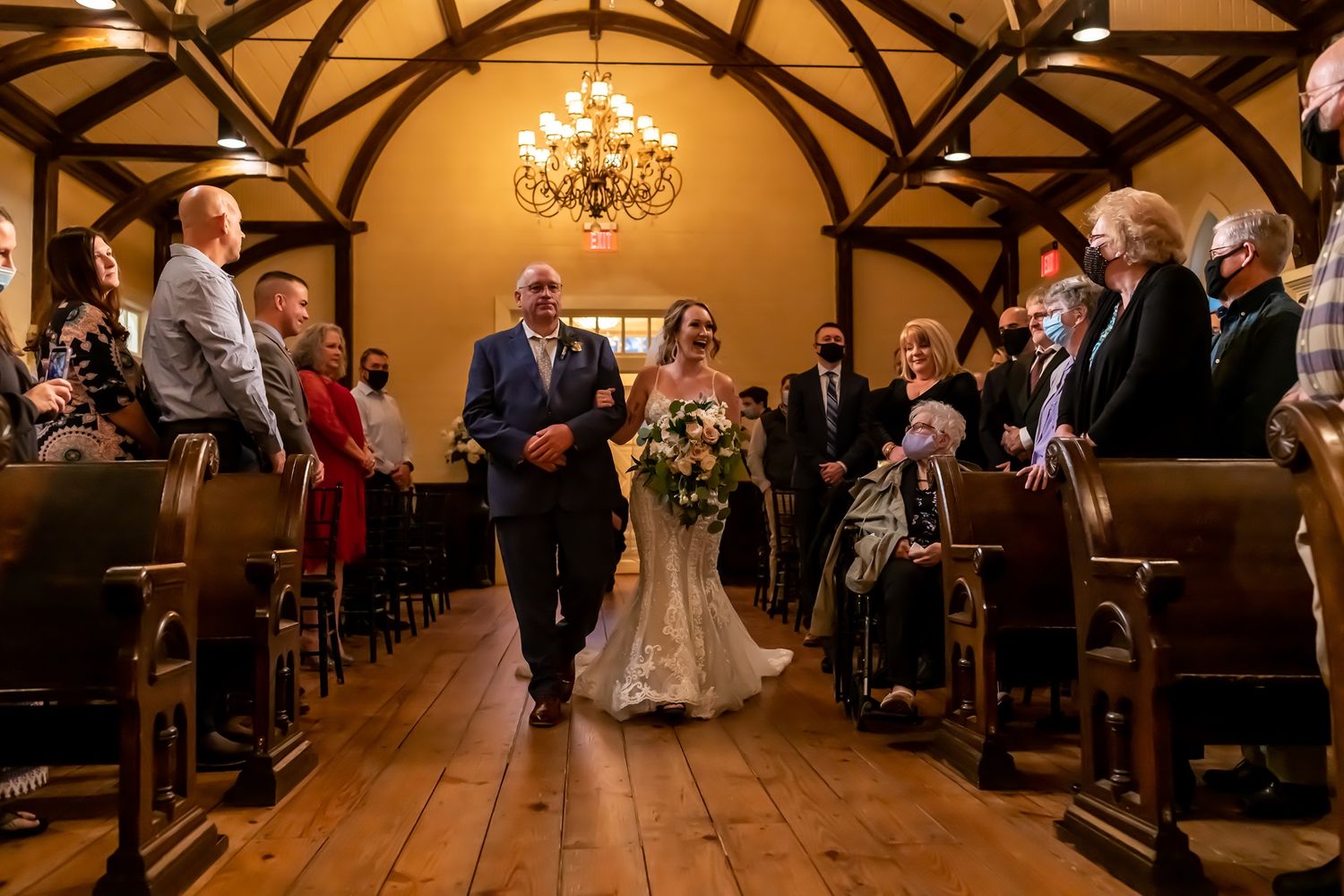 A wedding ceremony procession in a rustic wooden chapel with exposed beams and a warm chandelier overhead.