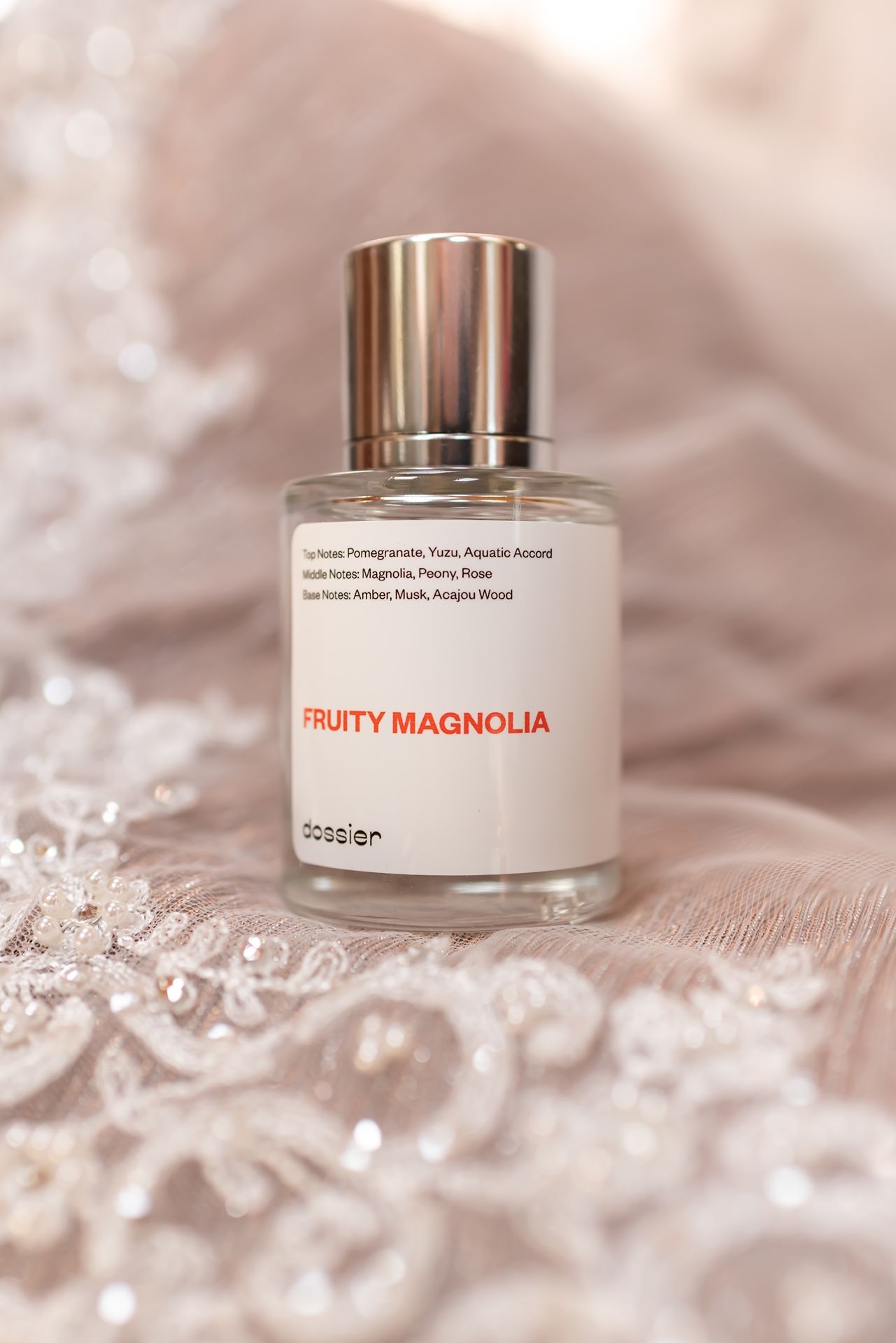 A bottle of Fruity Magnolia perfume with silver cap sits on delicate lace fabric with soft blurred background.