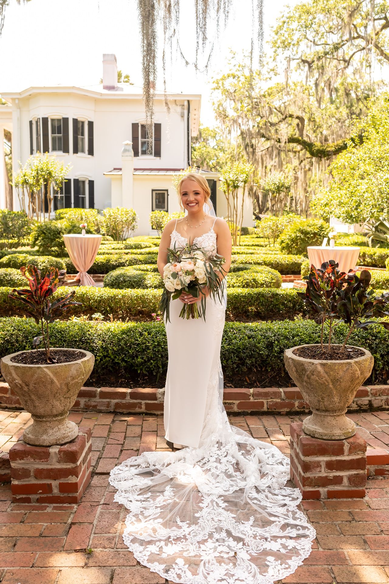 A bride poses in her lace wedding gown with a flowing train on a brick patio with garden planters.