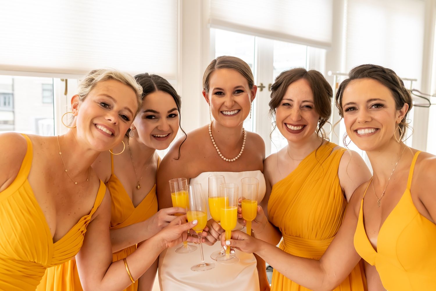 A group poses together while holding champagne glasses wearing gold bridesmaid dresses and a white wedding gown.