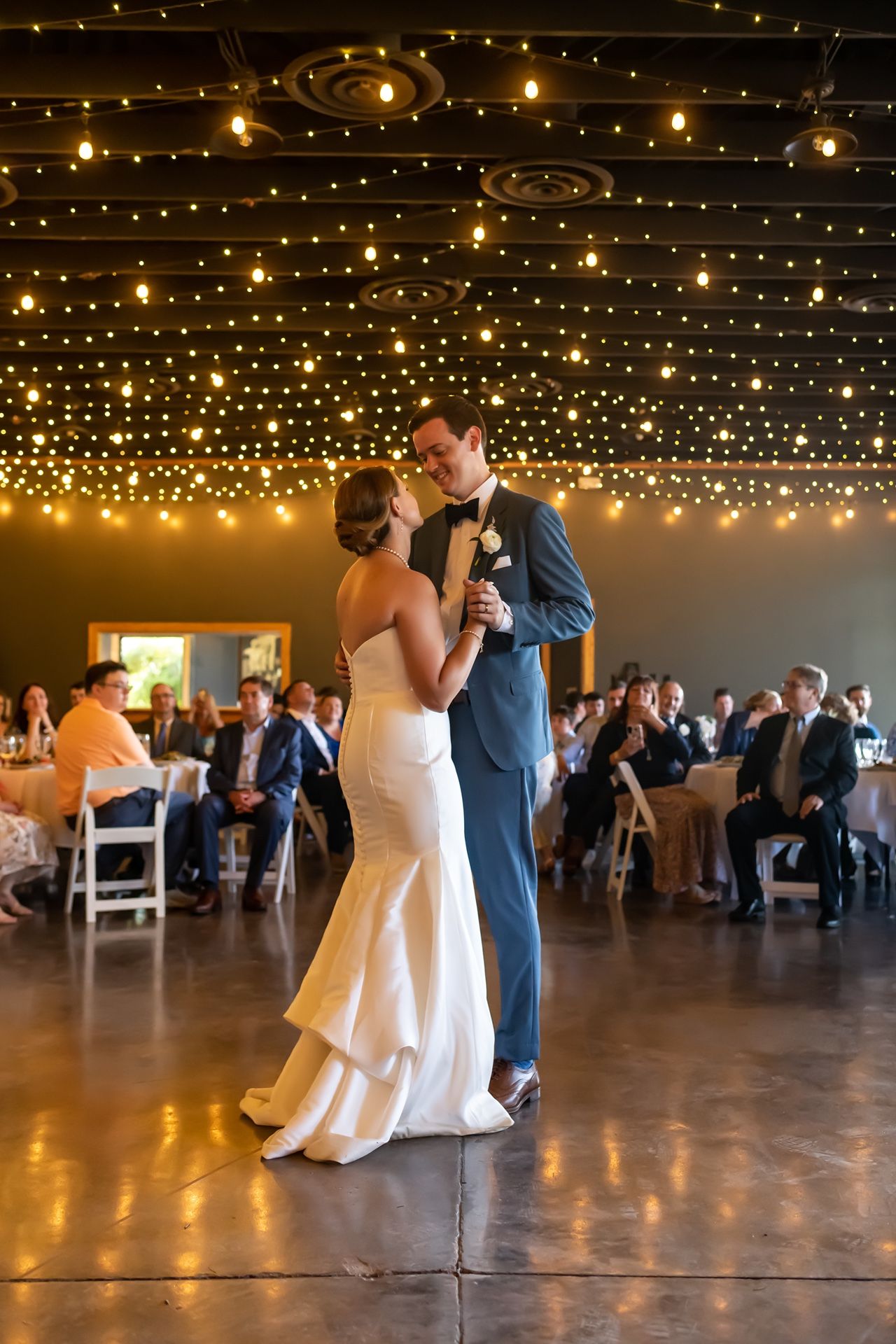 Couple sharing their first dance under twinkling string lights at an elegant indoor wedding reception.