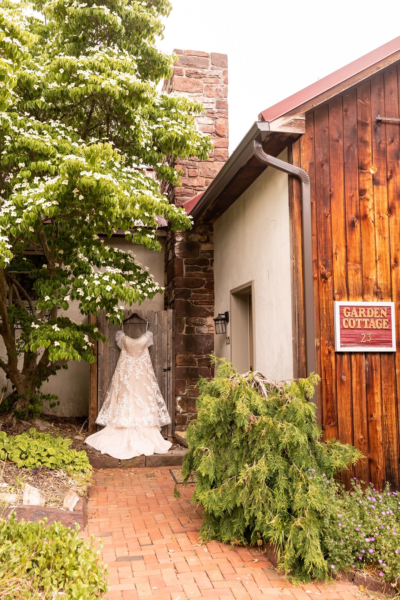 White wedding dress hanging on wood door in garden cottage pathway with red brick pavement and green foliage.