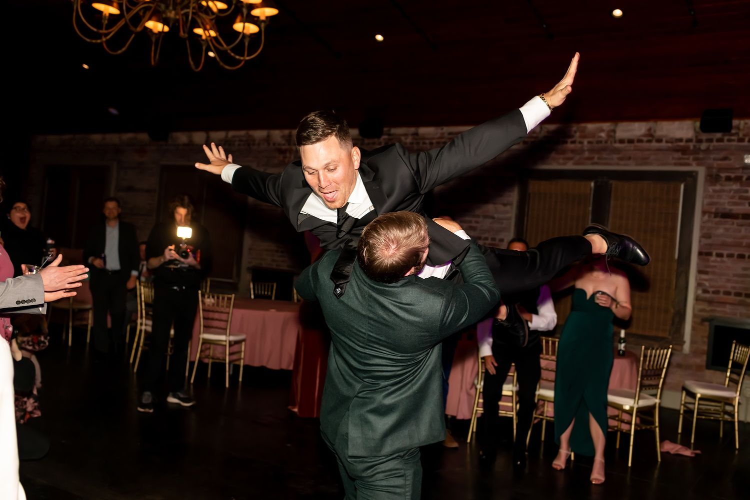 Two people celebrating at a wedding reception as one lifts the other high in the air on the dance floor.