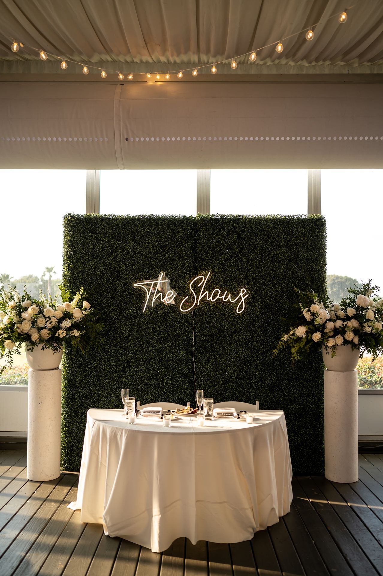A wedding sweetheart table setup with neon sign against a boxwood wall, white linens, and floral arrangements.