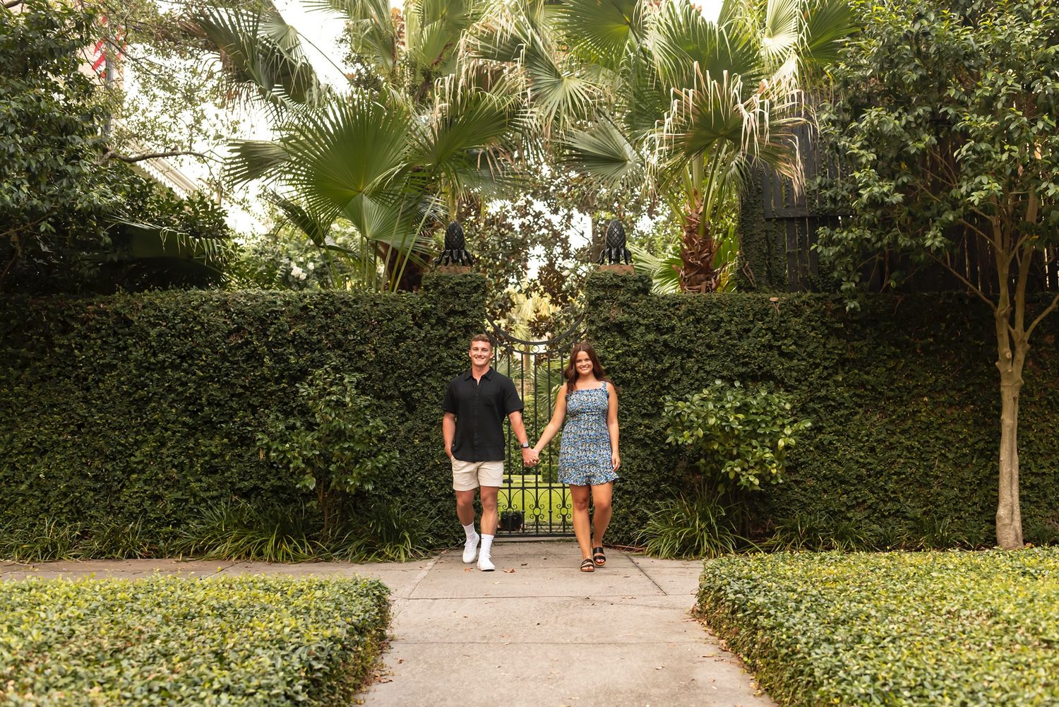 Two people walk together on a winding garden path with dense foliage and palm trees in the background.