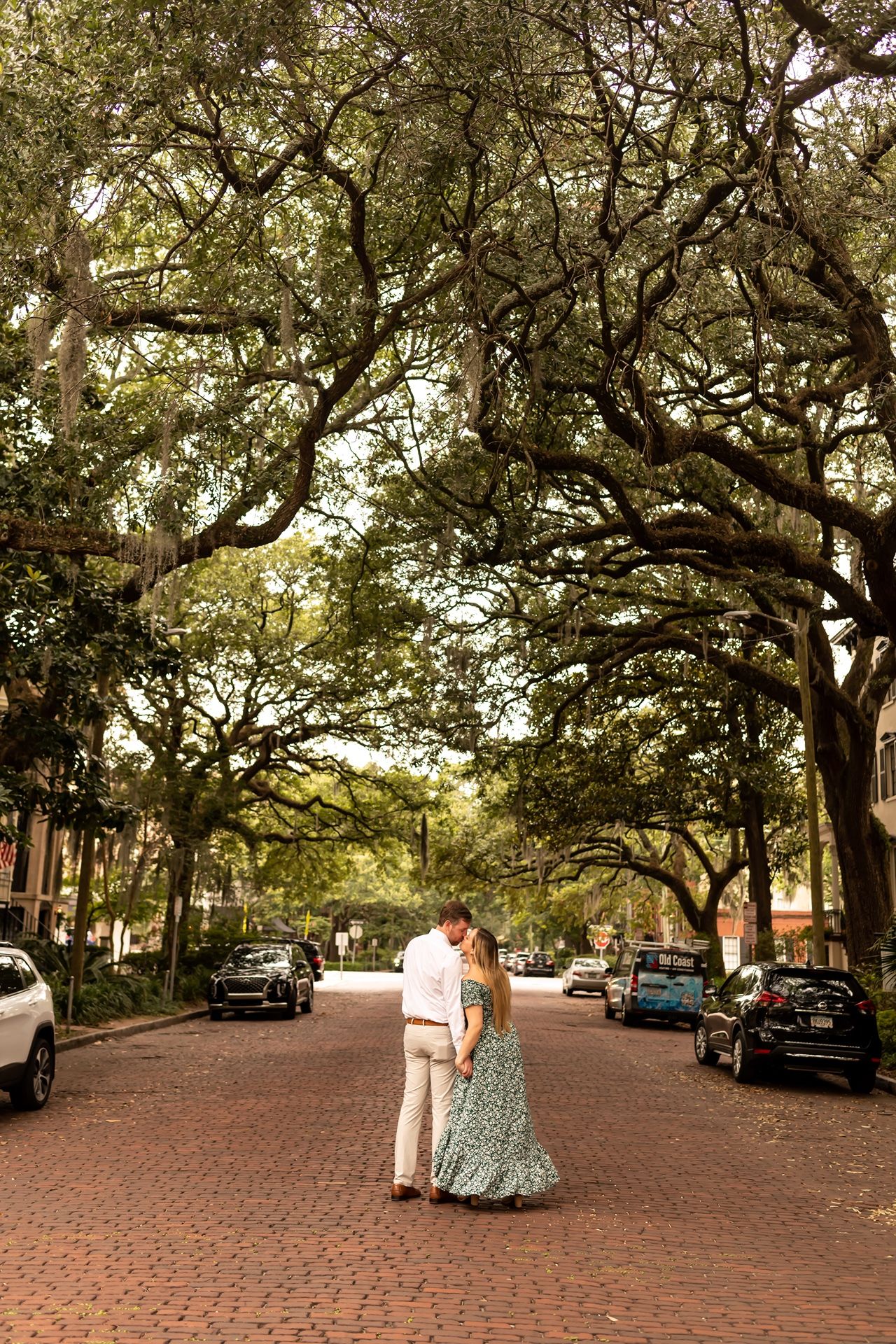 A romantic couple embraces on a brick-paved street lined with moss-draped oak trees and parked cars in Savannah, Georgia.