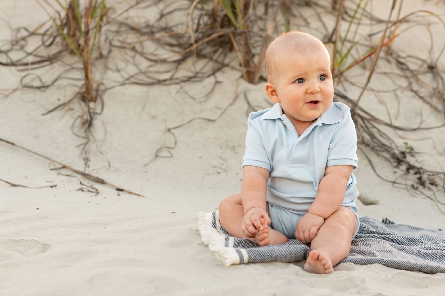 Baby in light blue onesie sits on sandy beach against weathered wooden fence background.