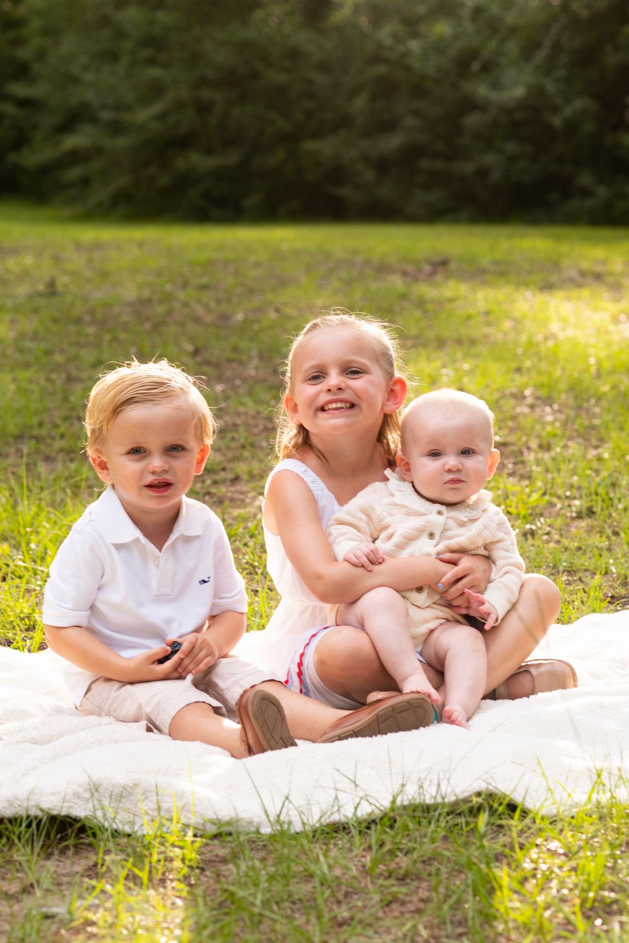 Three young children sitting on a white blanket in a sunny park, smiling for a family photo outdoors.