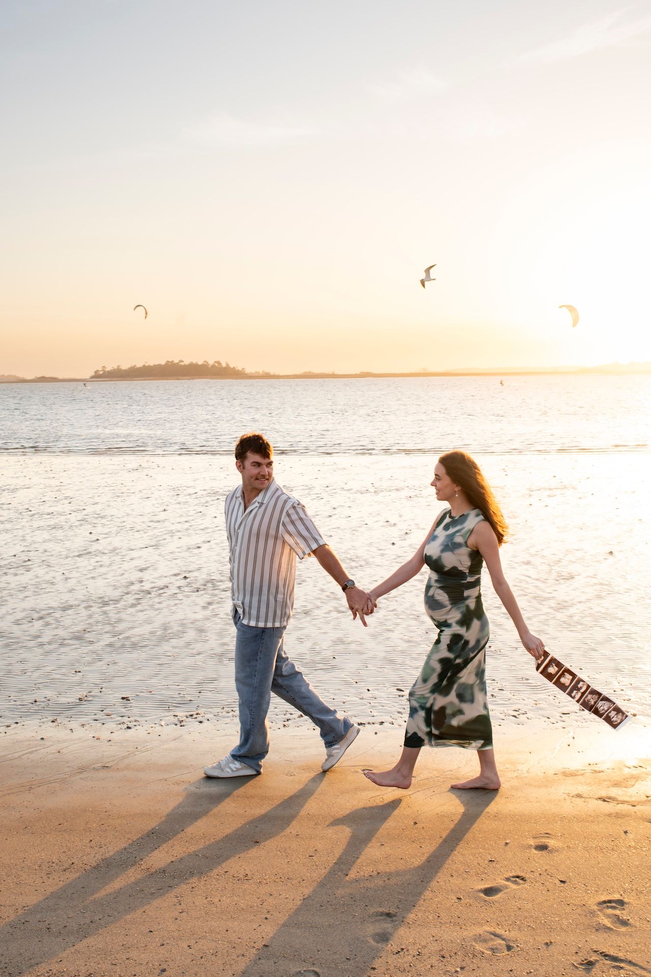 A couple dances playfully on a sandy beach at sunset casting long shadows on the sand as waves roll in.