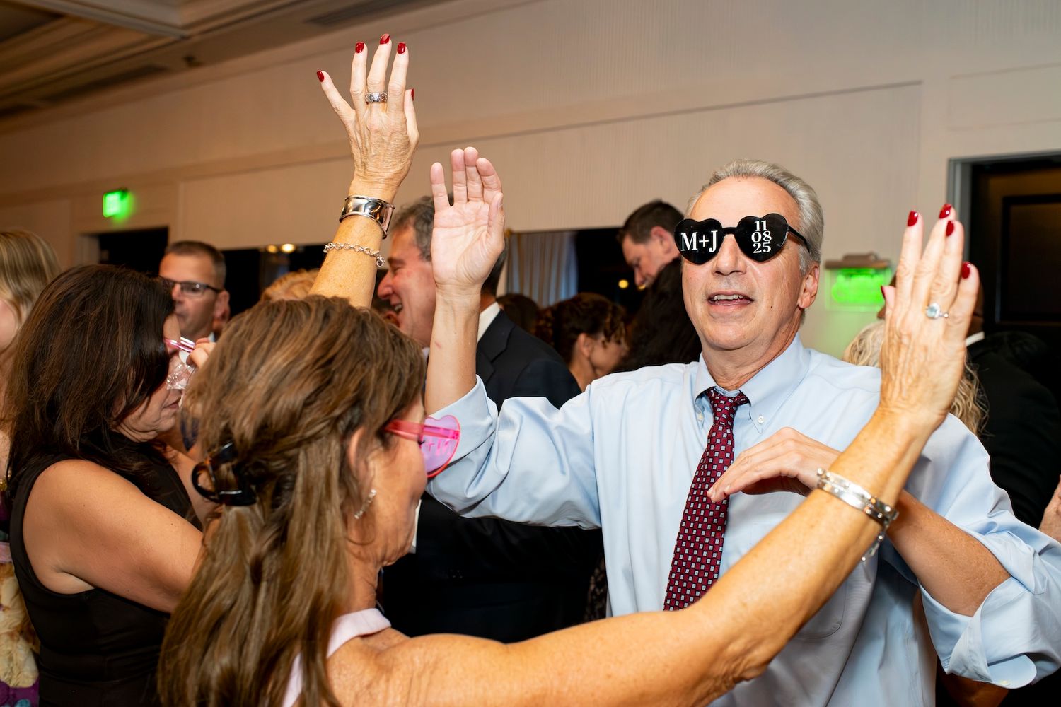 Group of wedding guests dancing with raised hands on a dimly lit dance floor during a celebration.