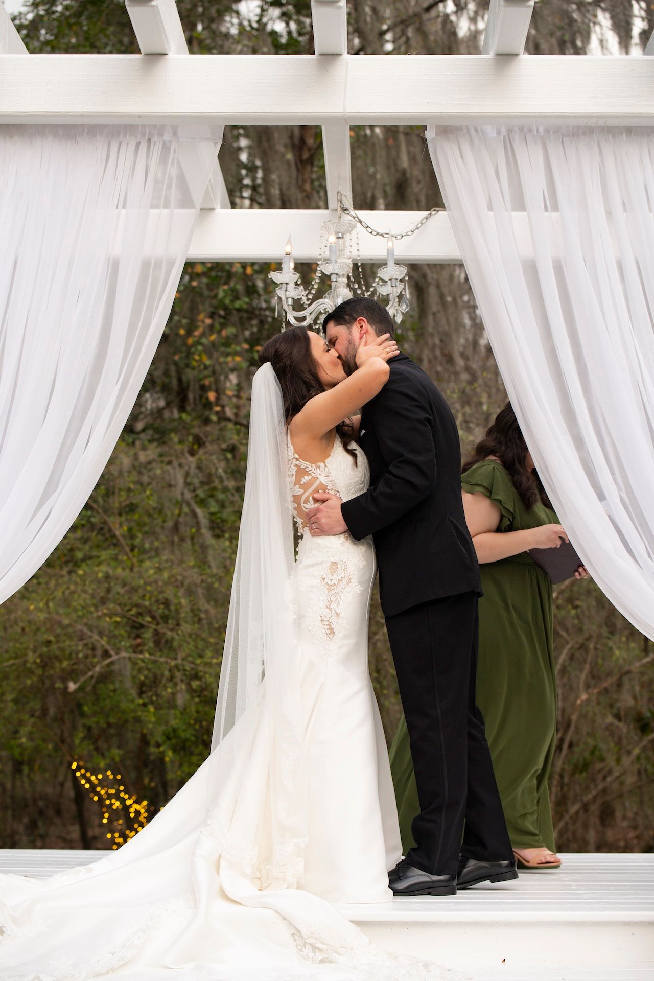 Bride and groom share first kiss under white draped pergola with chandelier at outdoor wedding ceremony.