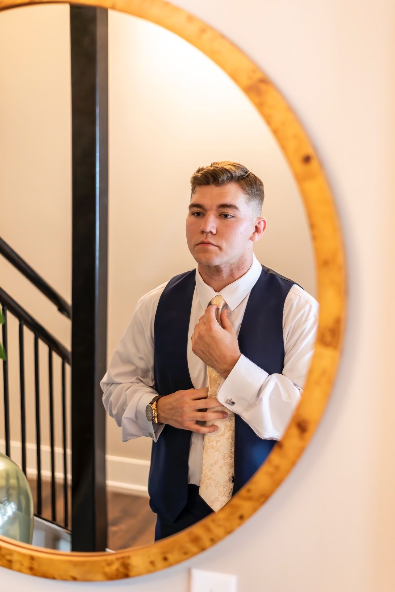 A groom adjusts his champagne-colored tie while looking in a round wooden-framed mirror.