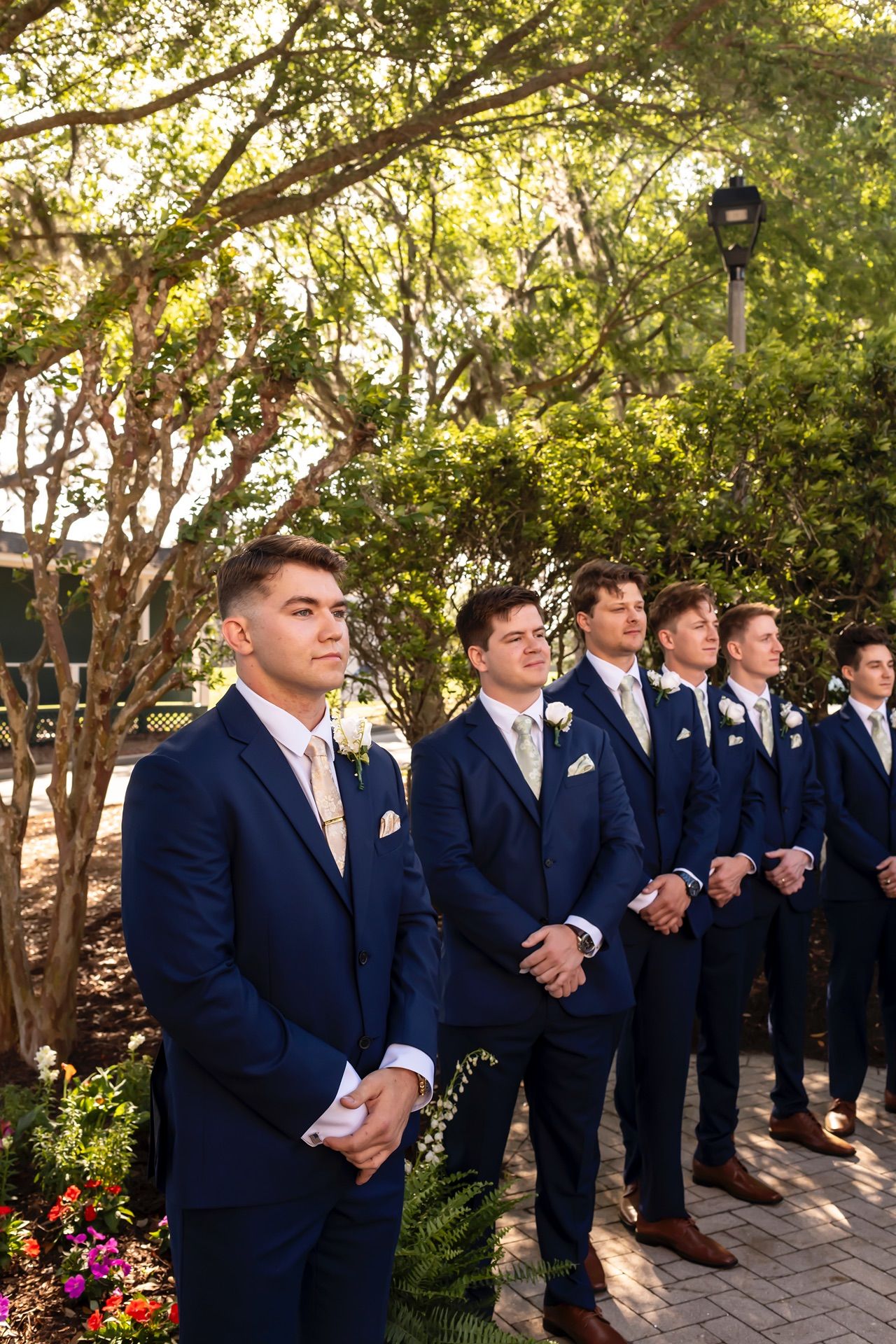 Groomsmen in navy suits stand in a line outdoors under sunlit trees during a formal wedding ceremony.